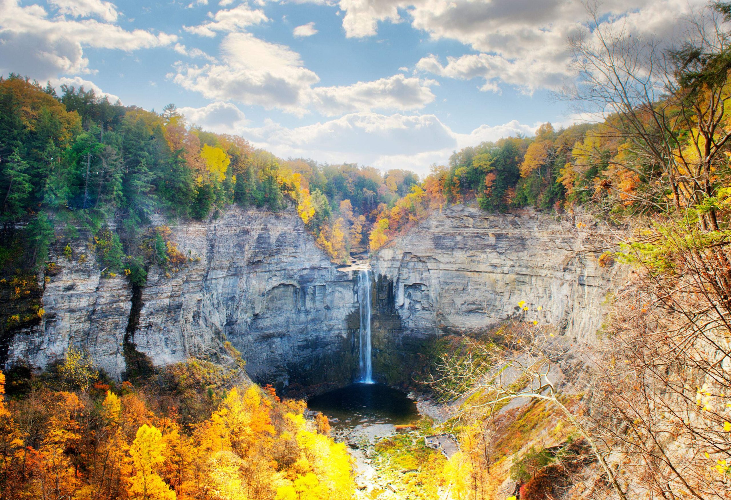 A spectacular waterfall with clear free-falling water between rocky mountains with countless autumn trees atop and around.