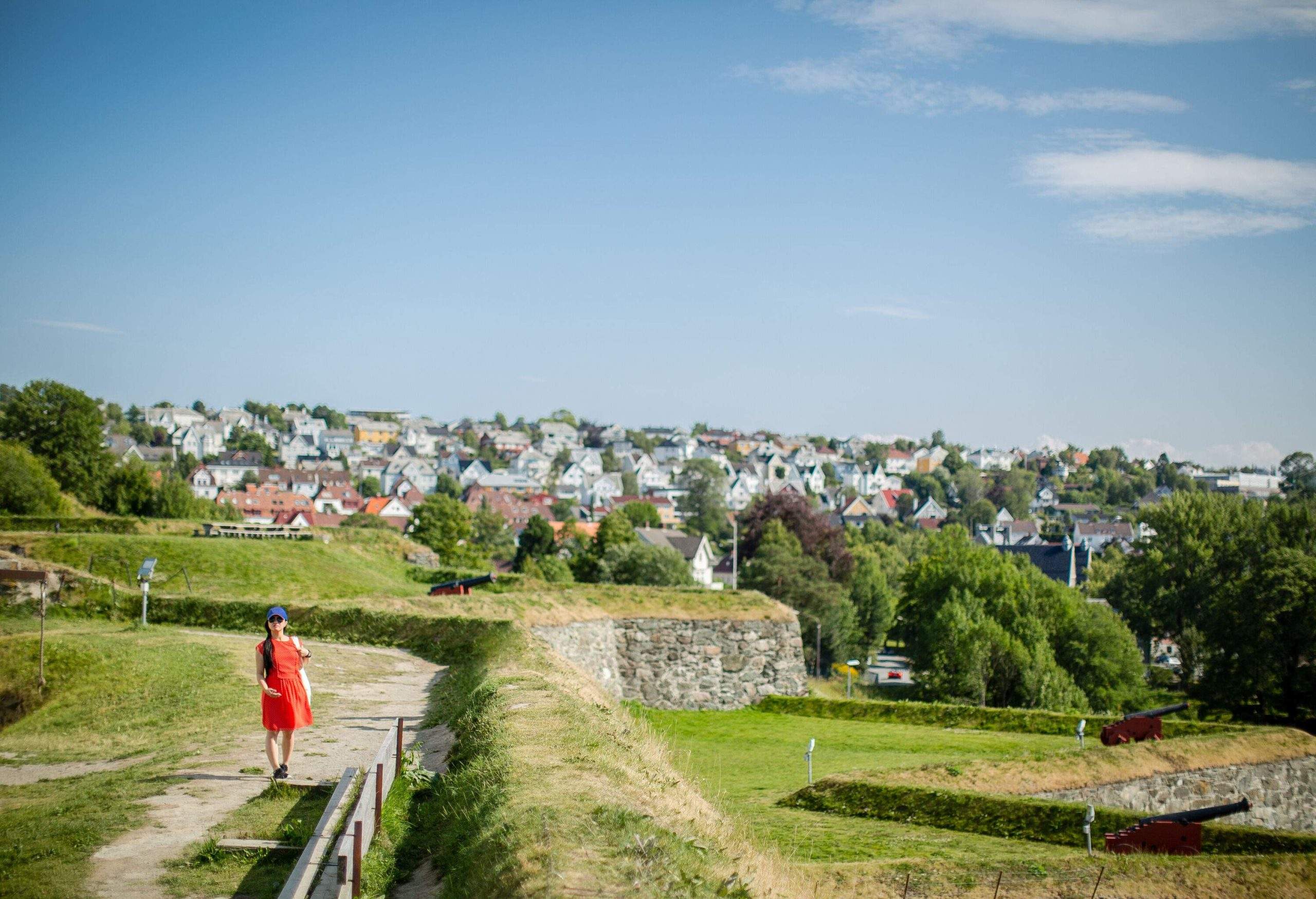A woman in a red dress stands on the top of a fortress overlooking a cluster of buildings in the distance.