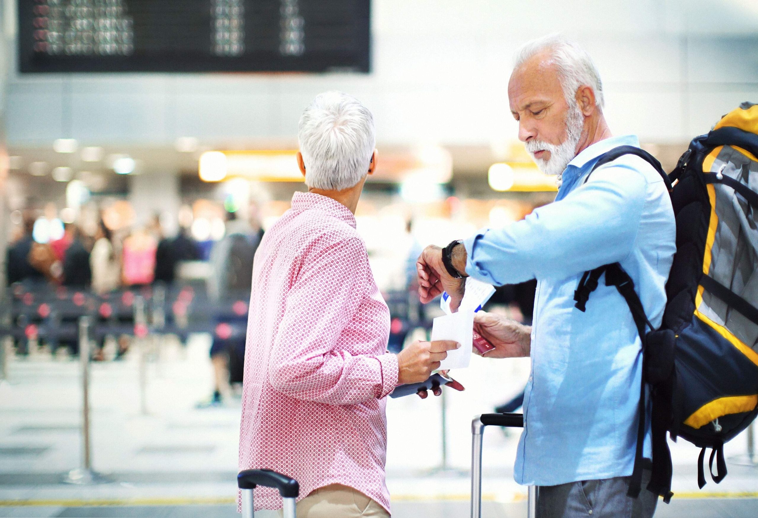 Closeup side view of senior couple at an airport waiting for their flight which has been delayed. They are both holding their passports and tickets. The lady is looking at the arrival departure board...NOTE: tickets are generic with no data