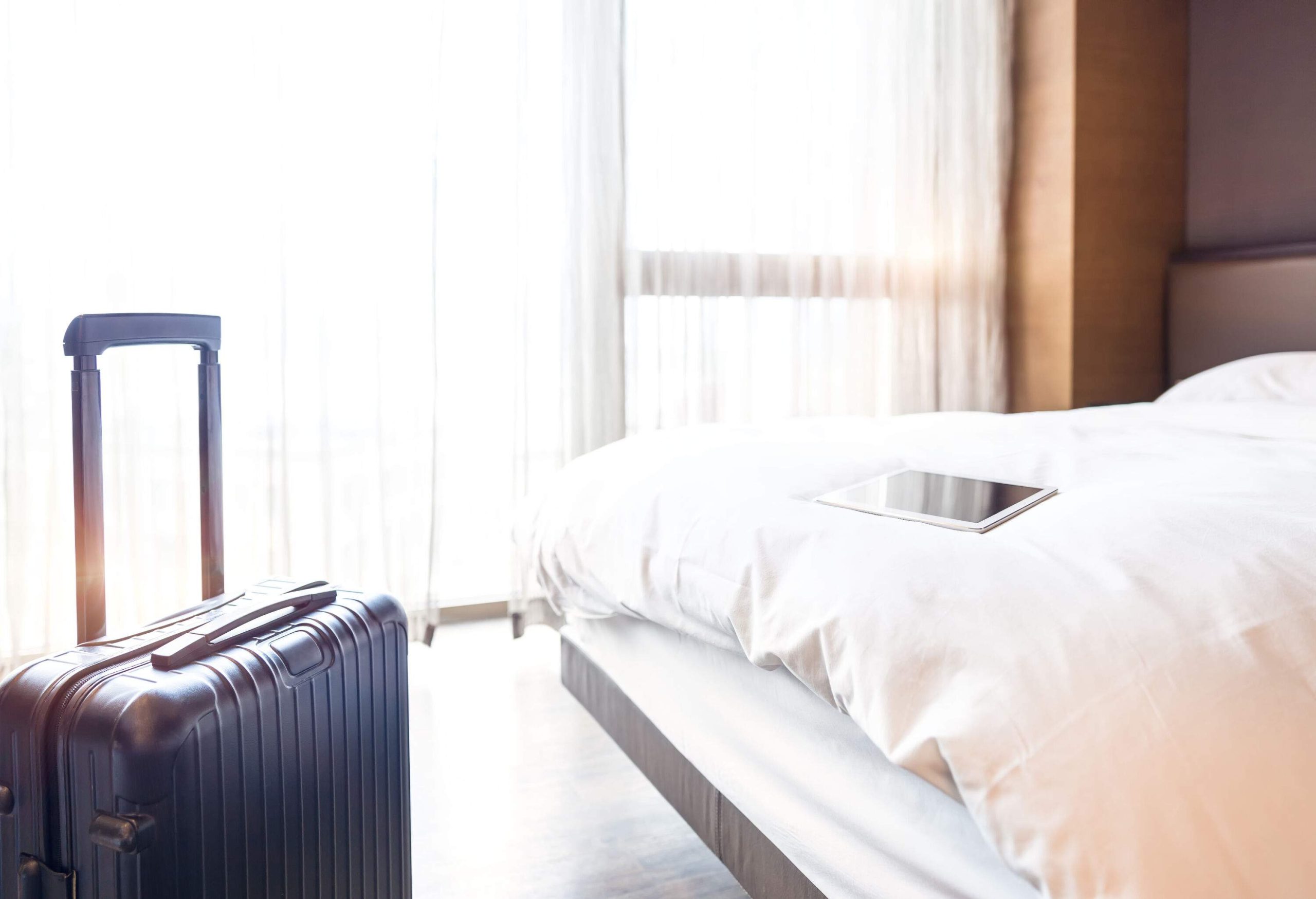 A hotel room with a tablet on a bed beside a black luggage.