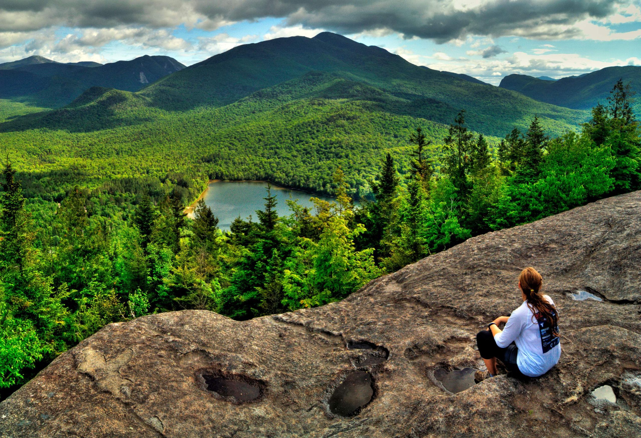 A person sits on a cliff overlooking the glorious verdant forested hills and a lake in the centre.