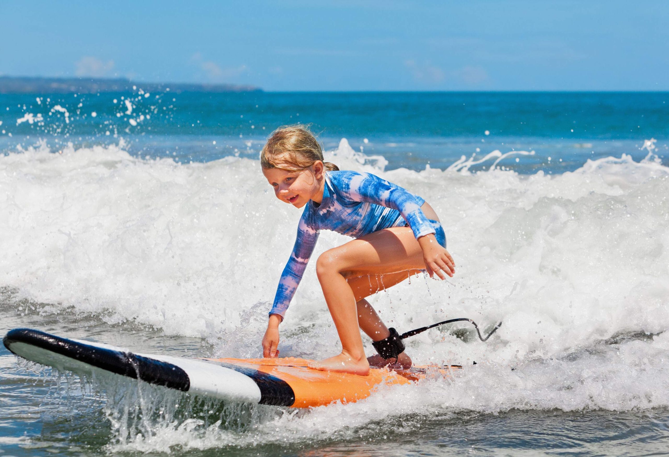 A young blonde girl smiles as she balances on a surfboard with crashing waves behind her.