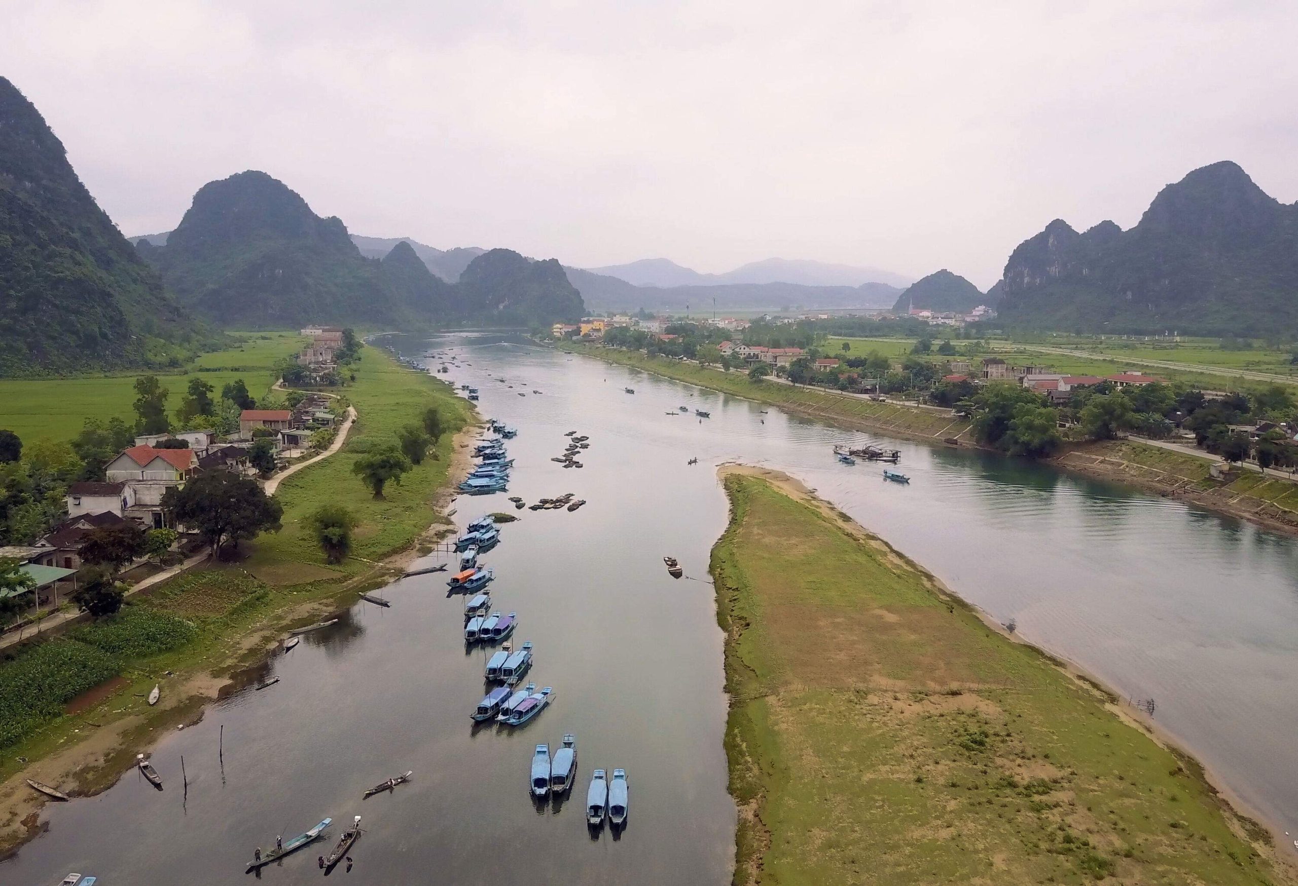 Numerous boats on the broad river bisecting the flat vegetated land between the steep mountains.