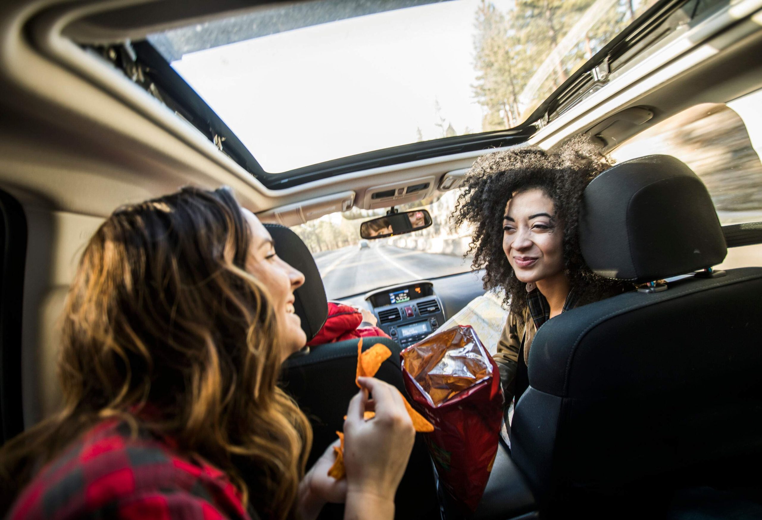 Two female friends having a conversation inside a car while eating chips.
