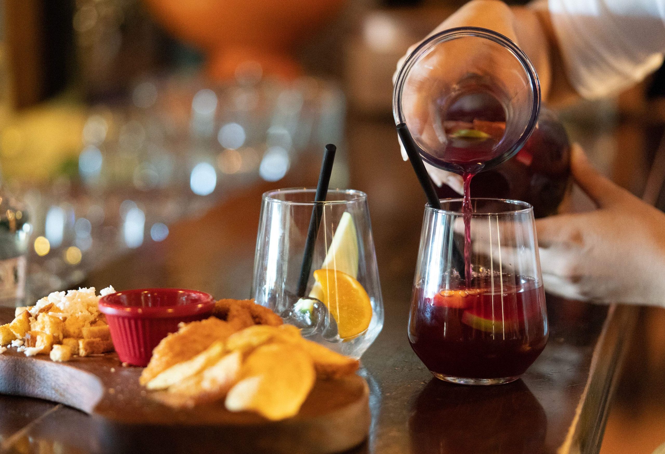 A pair of hands pouring a drink from a glass container into a glass with lemon slices served with bread, meat, and sauce.