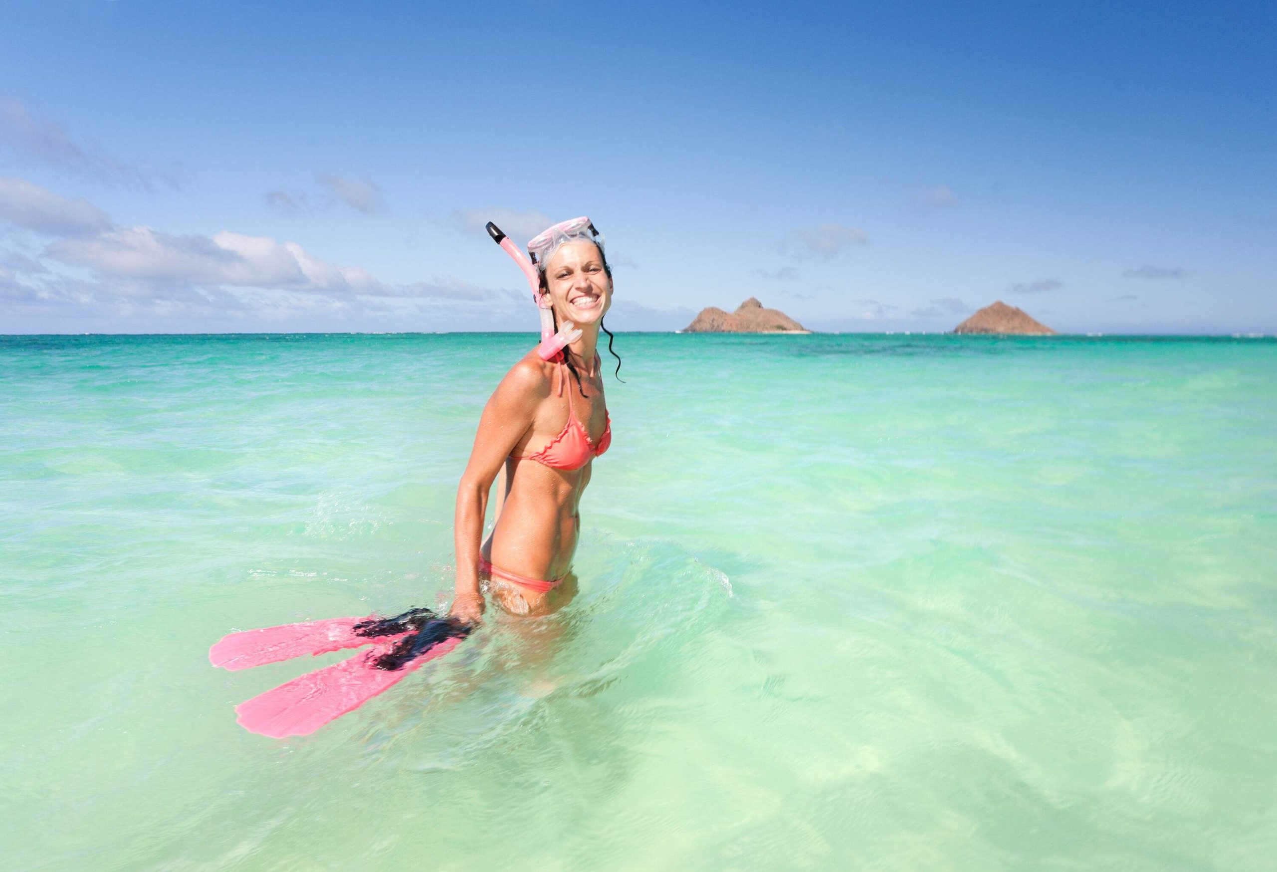 A woman in a bikini with her snorkel gear stands on the sea's shallow waters with two protruding rock mountains in the background.
