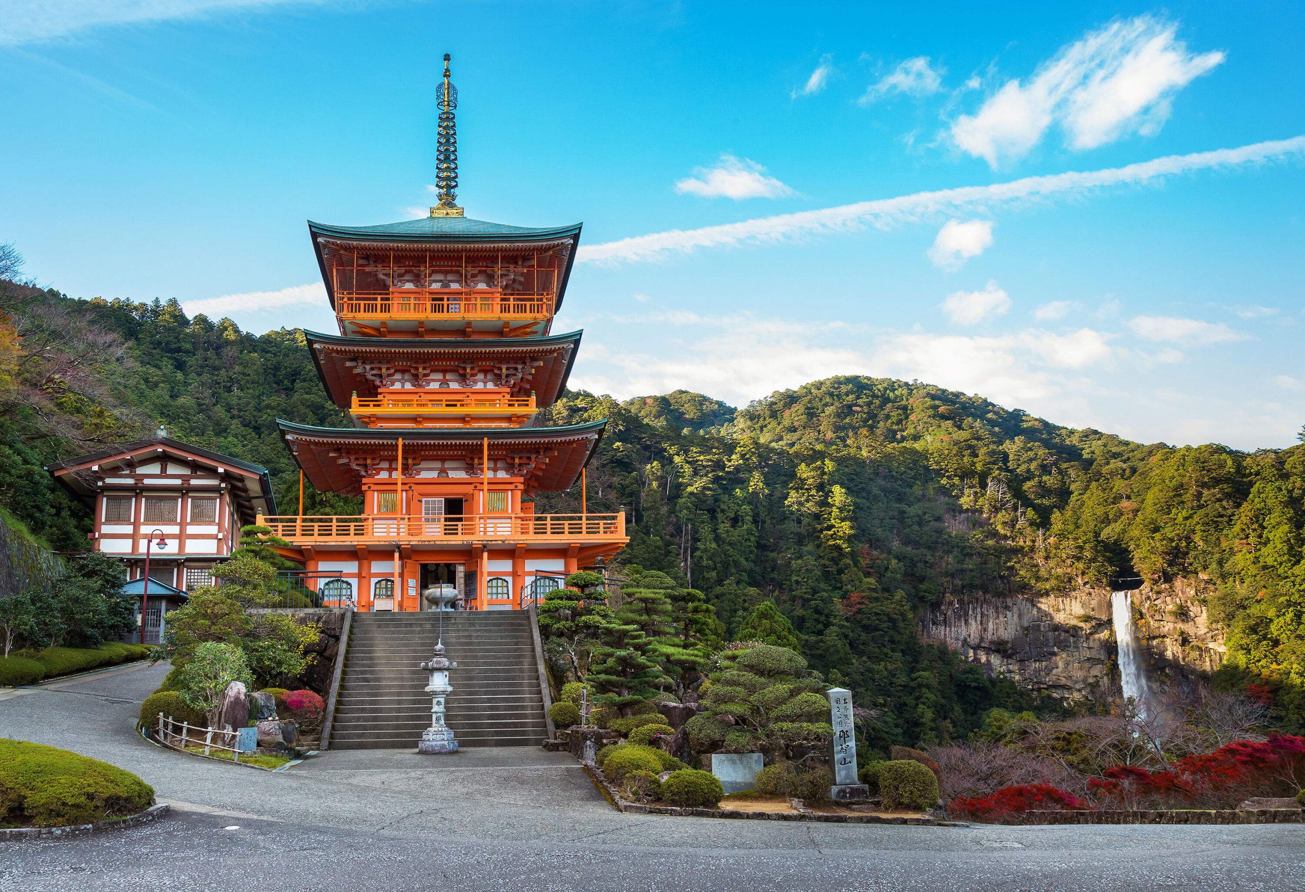 Stunning pagoda temple set against a backdrop of forested mountain range and cascading waterfalls beneath a serene cloudy blue sky.