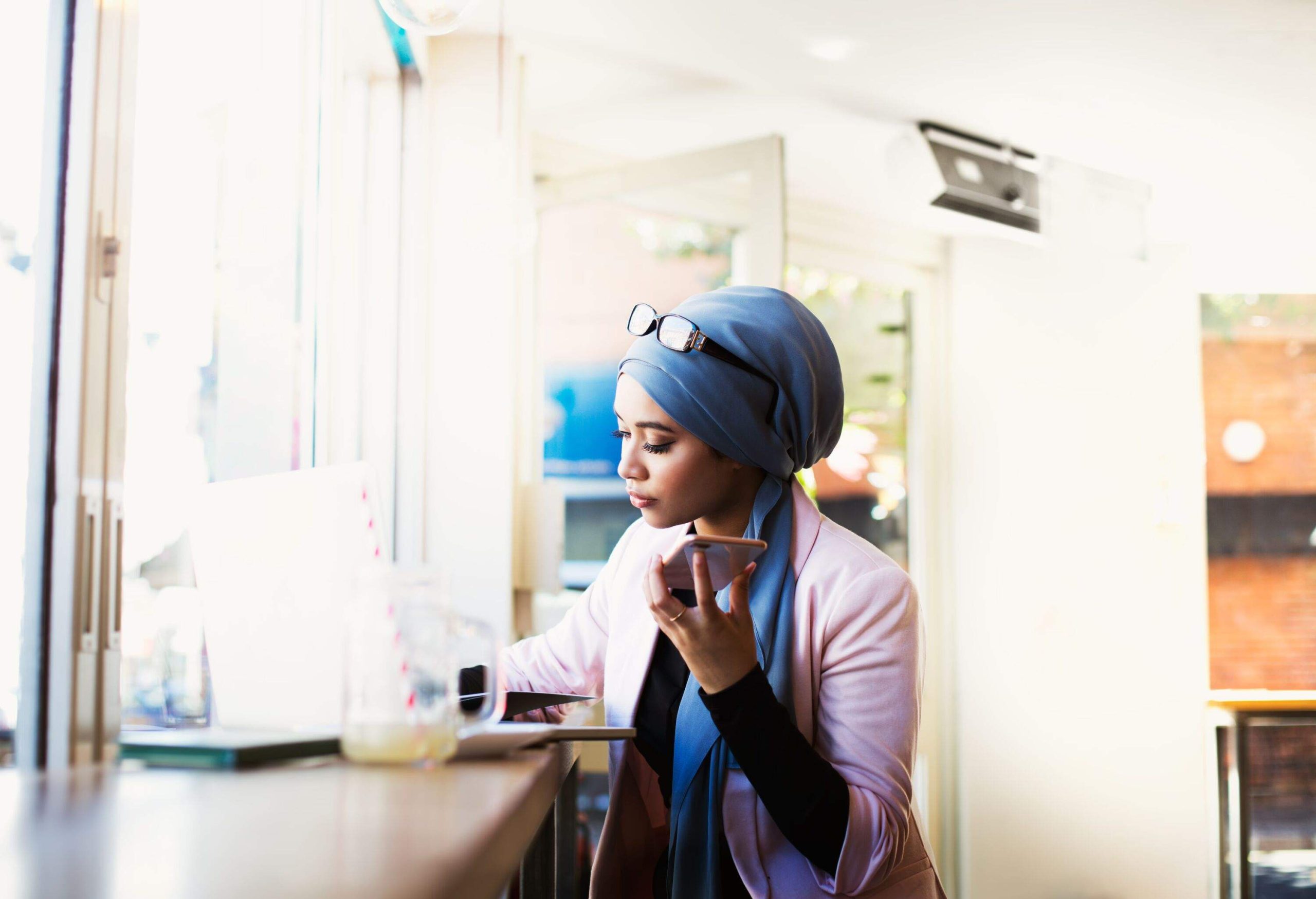 Woman wearing hijab working at laptop and looking at papers in coffee shop