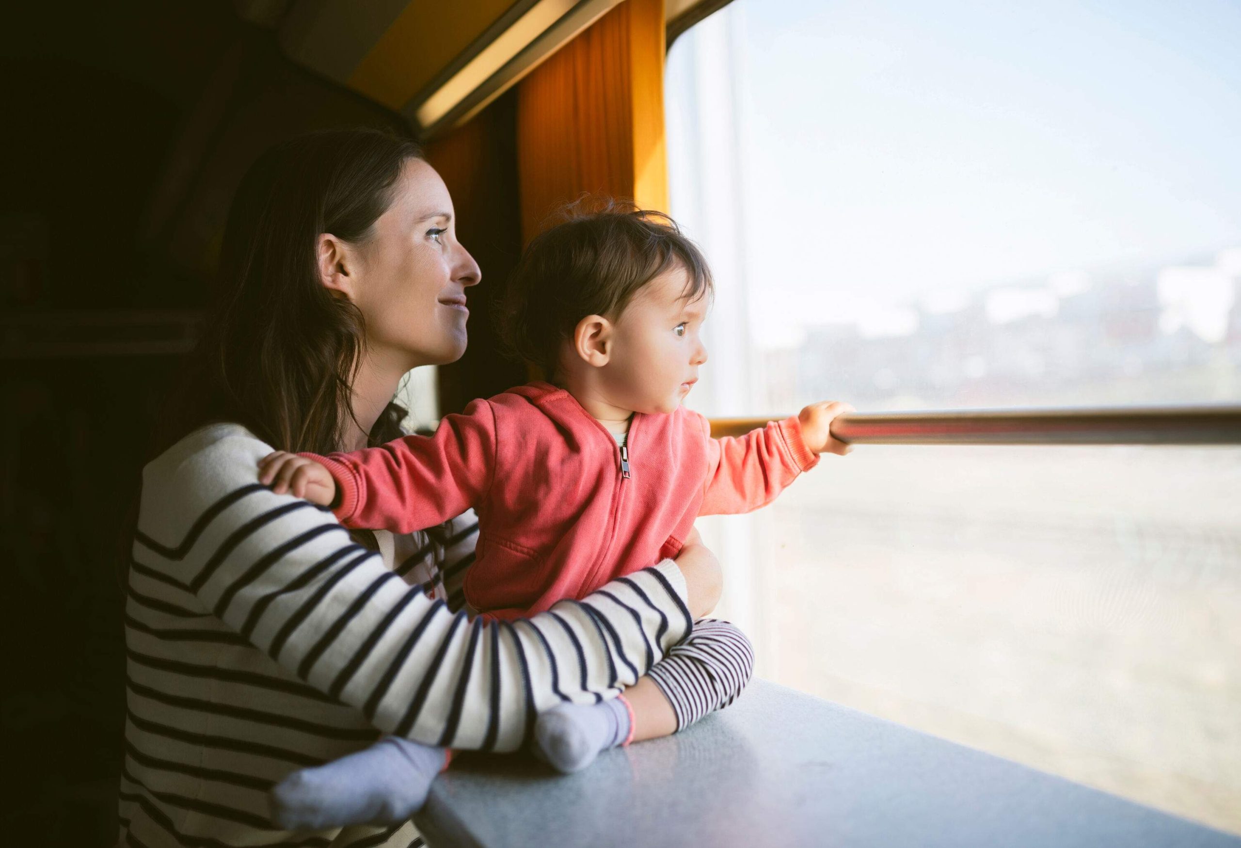 A mother in a striped sweater hugs a baby as they both look out the train's window.