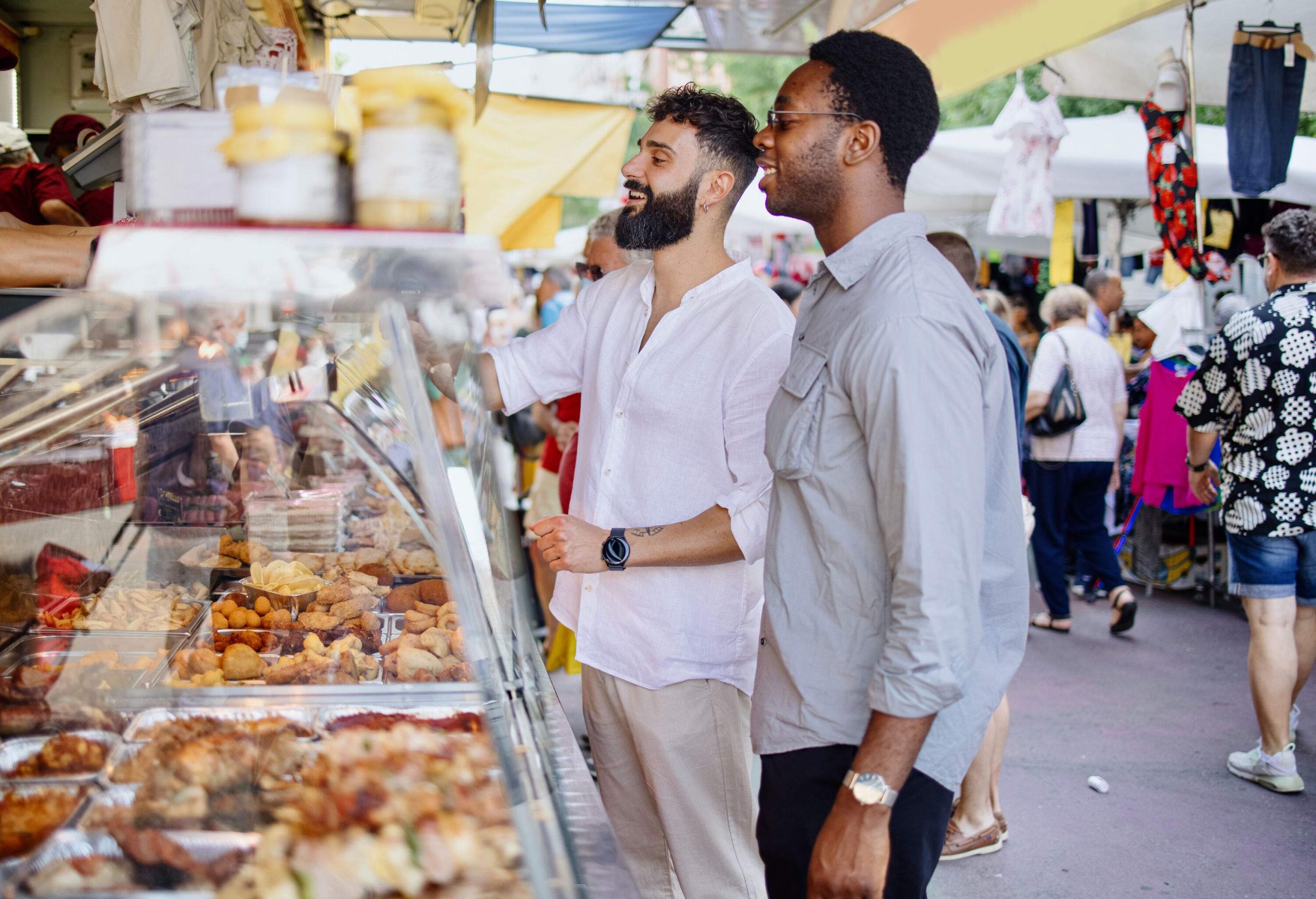 In the midst of a vibrant market, a cheerful couple of friends can be seen joyfully purchasing food from a nearby stall as people leisurely stroll by in the background.