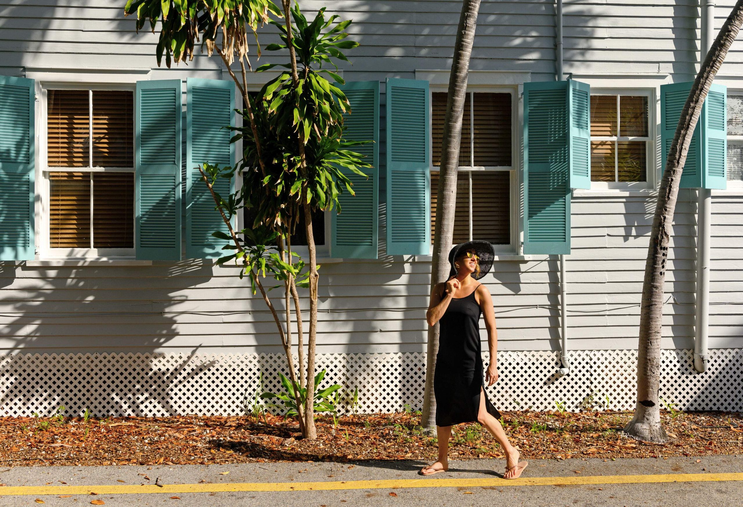 A woman in a black dress and a wide-brimmed hat strolls across a sidewalk beside a white residence.