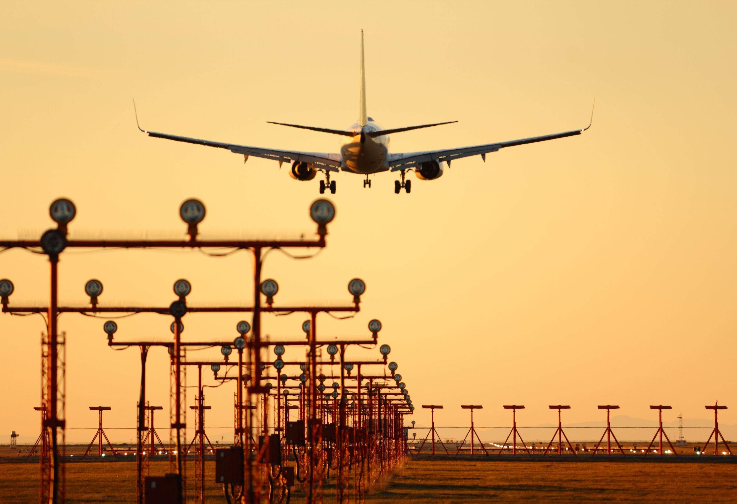 An airplane, landing gear extended, approaches a runway as the sun sets majestically, with vibrant runway markers guiding its descent in the foreground.