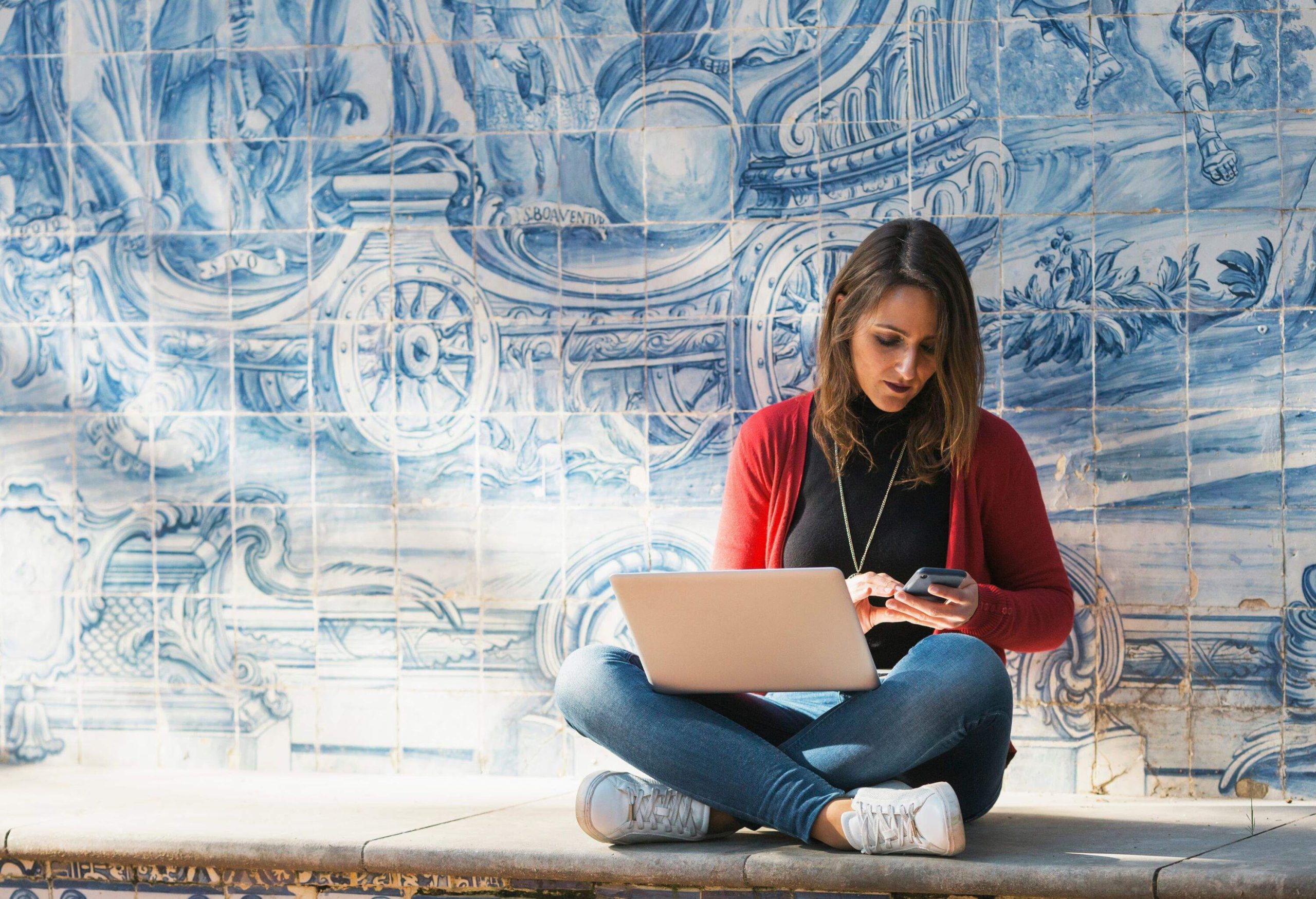 Woman sitting by a wall covered in azulejos working on her laptop and looking at her mobile phone