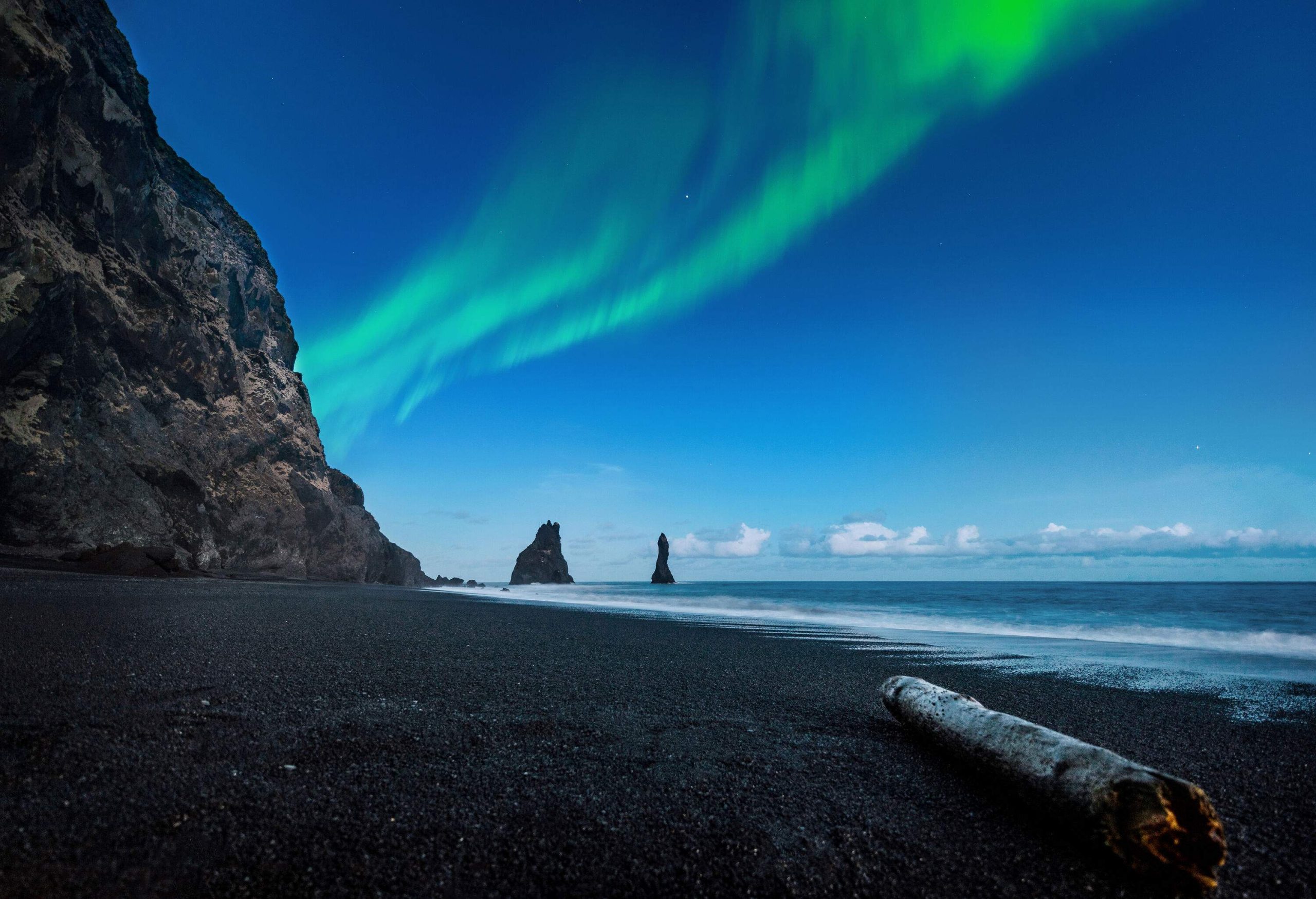A spectacular display of the northern lights fills the night sky above a peaceful beach with a sheer rock formation to the side.