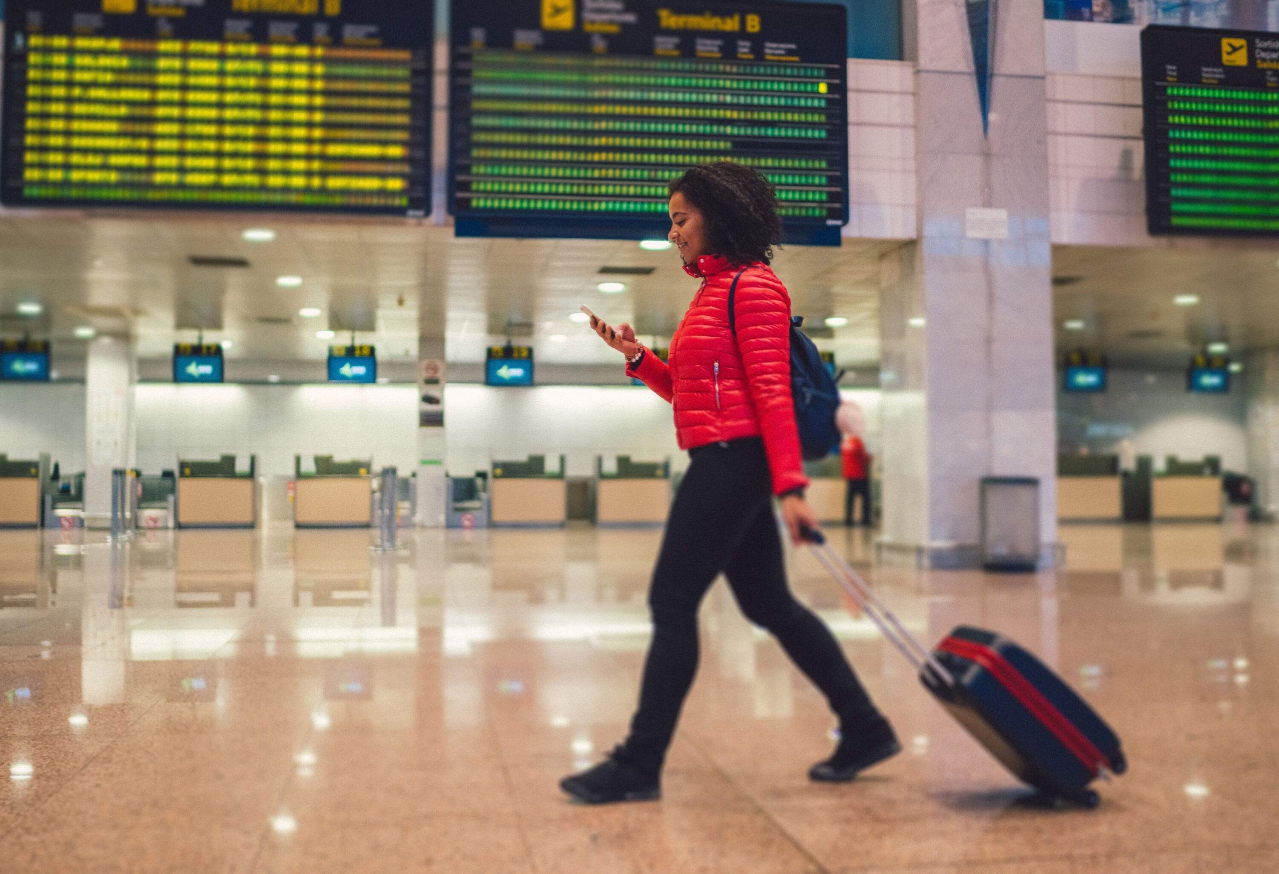 A woman in a red jacket checking her phone while hauling her luggage through the airport.