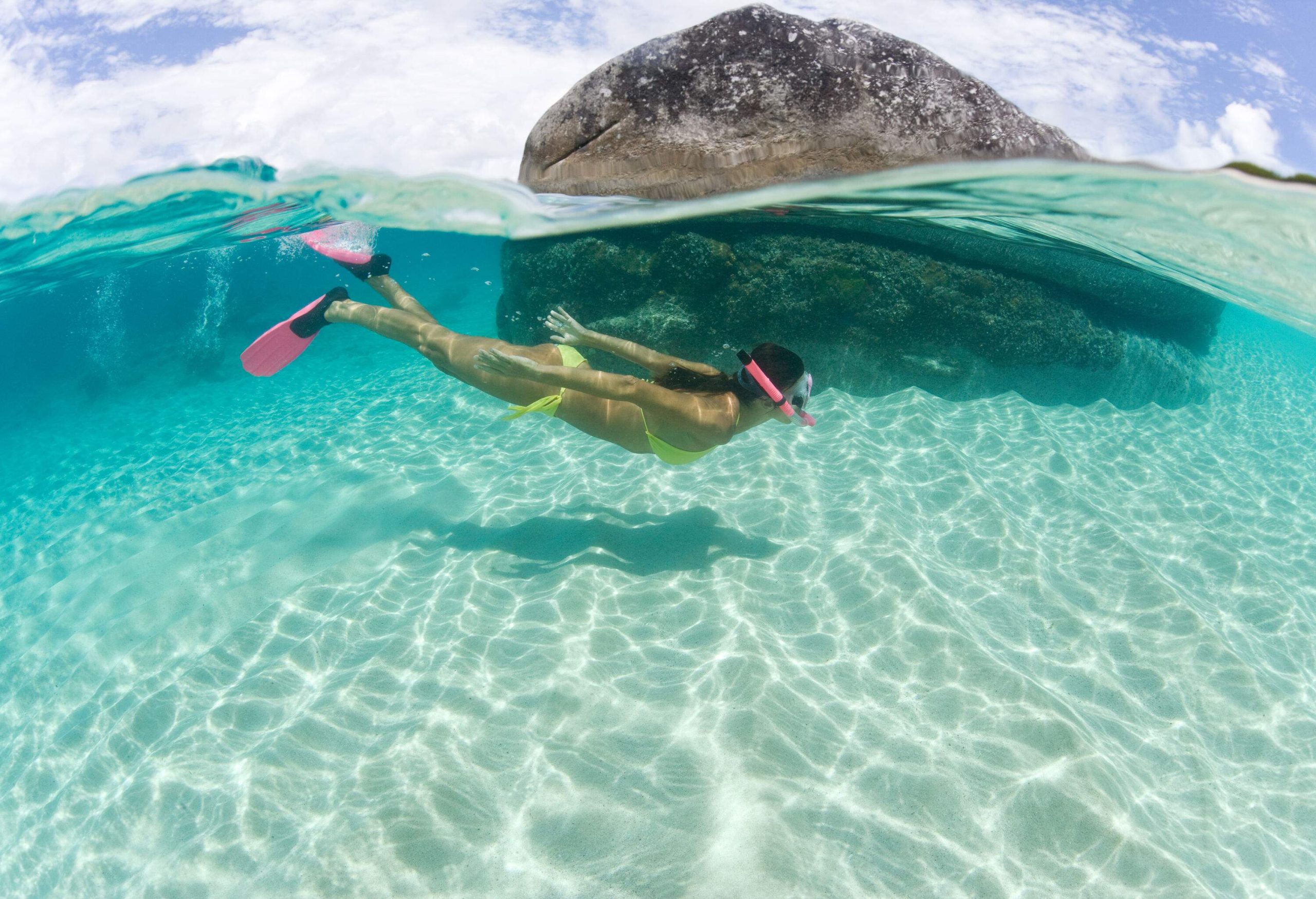 A woman in a bright green bikini driving around a huge sunken boulder in a crystal-clear sea.