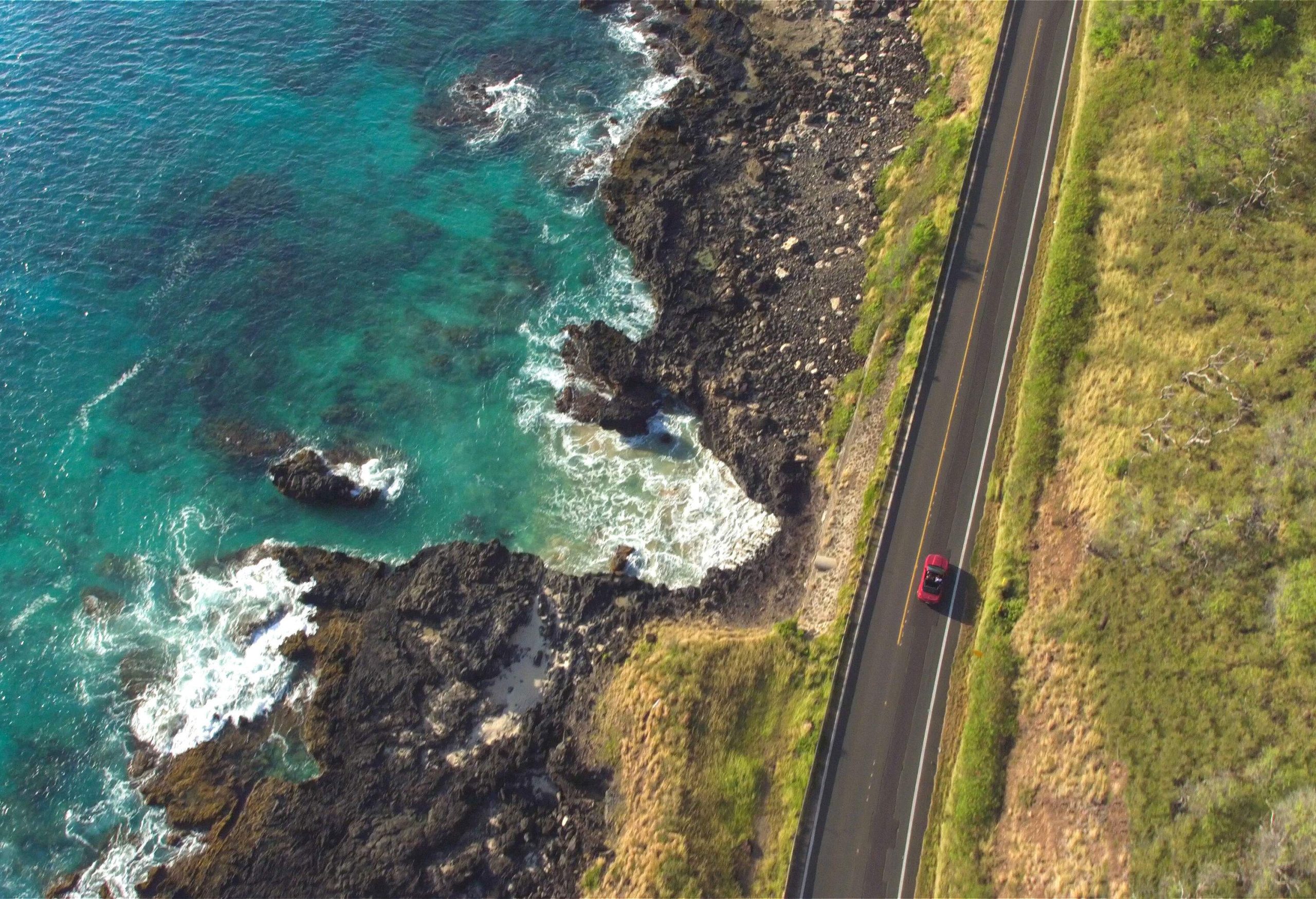 A car travels on a coastal road along a rocky beach with crashing waves.