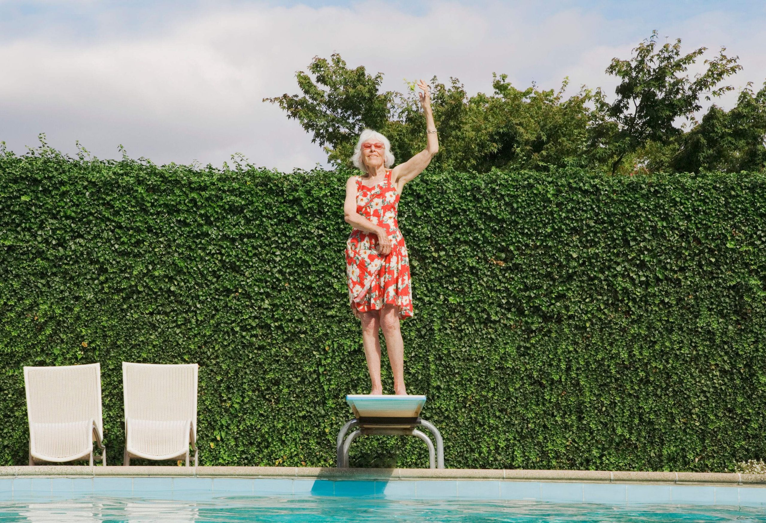 A senior woman in a floral dress is standing on a diving board above the pool against a green leaf wall.