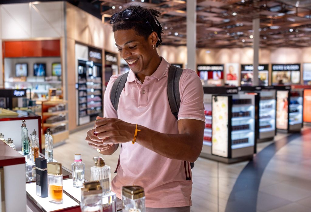 A side-view shot of a young man wearing casual clothing exploring a duty free shop in an airport in Toulouse, France before catching his flight. The man is smelling aftershave.