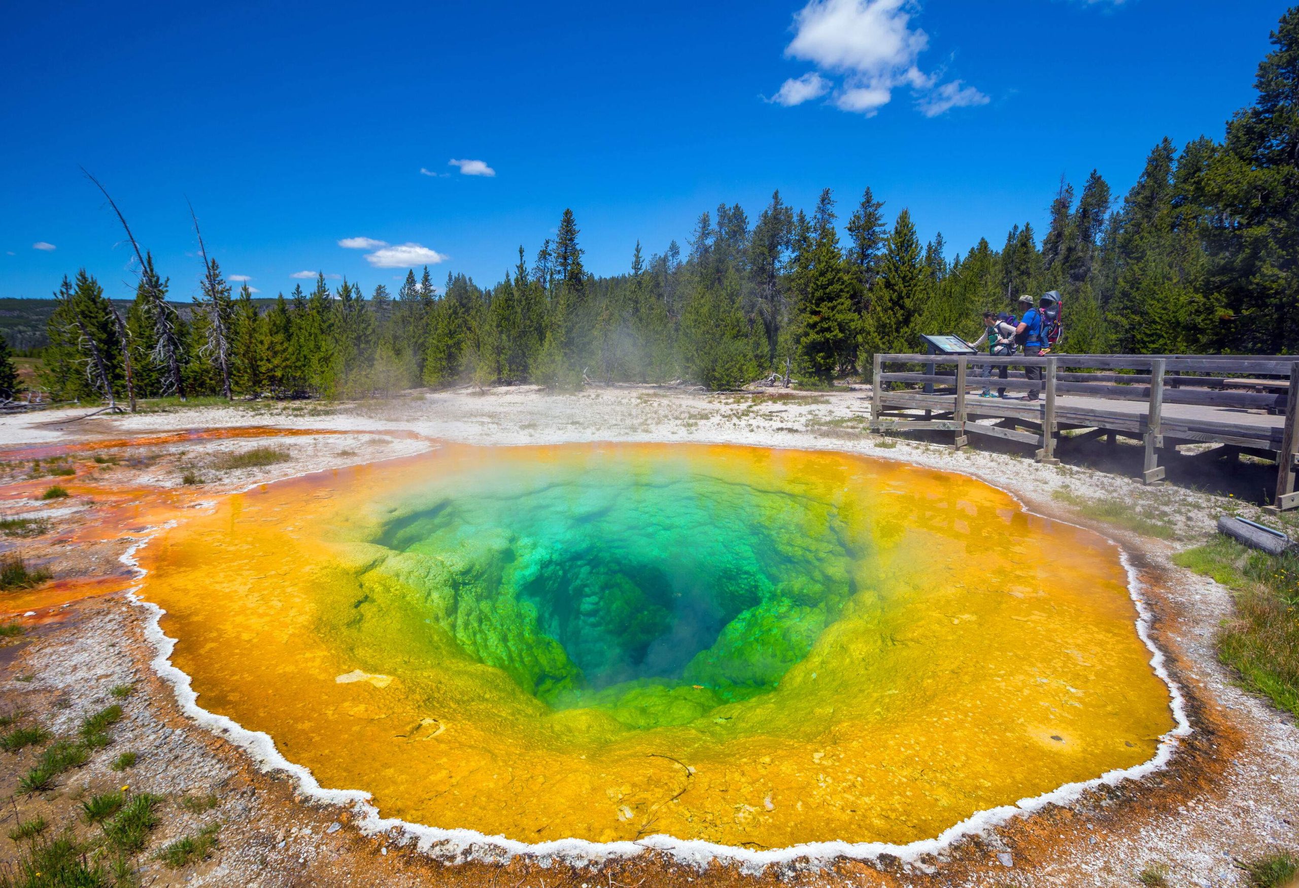 Two individuals are on a viewing deck looking at a hot spring with a distinct colour pool due to bacteria.
