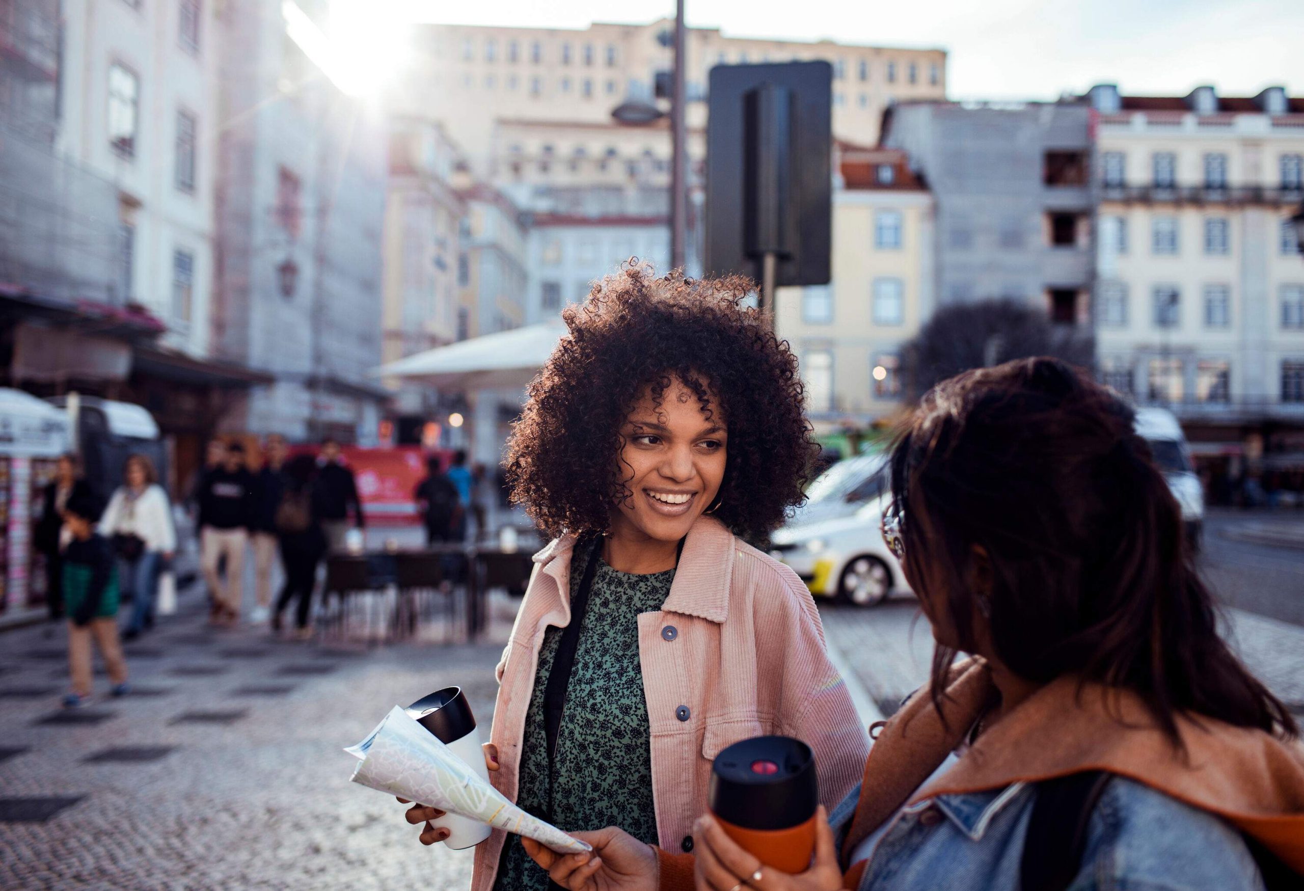 A curly-haired woman smiles at a friend while holding a cup and a map.