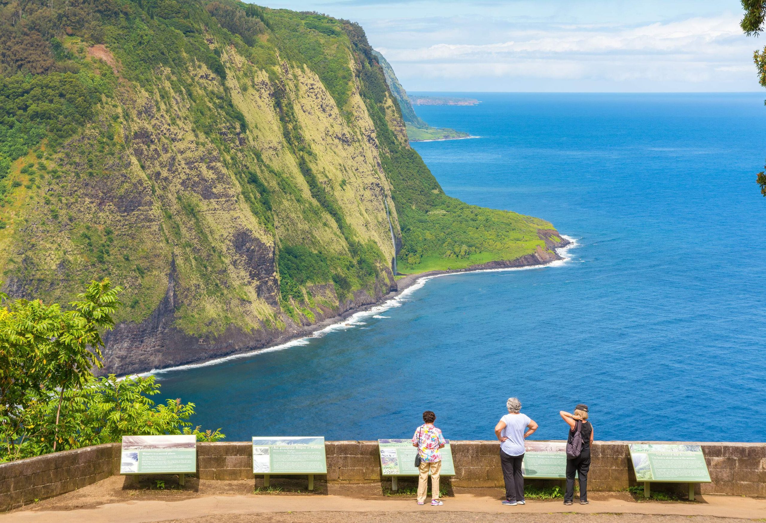 Three individuals on a viewing deck overlooking the forested cliff surrounded by the tranquil blue sea.