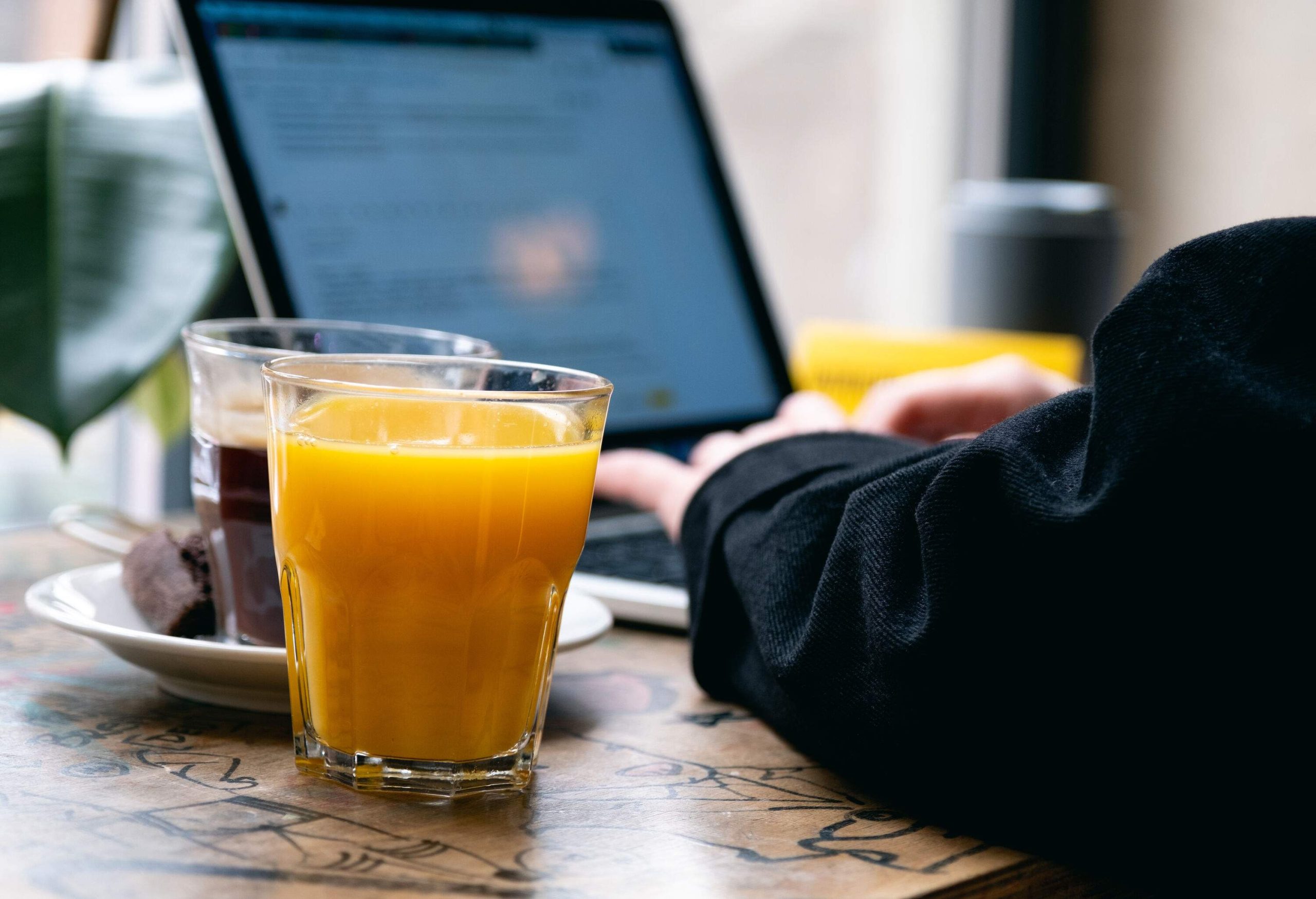 Focusing intently on their laptop, a person works diligently while a steaming mug of black coffee and a refreshing glass of orange juice sit nearby on the table.