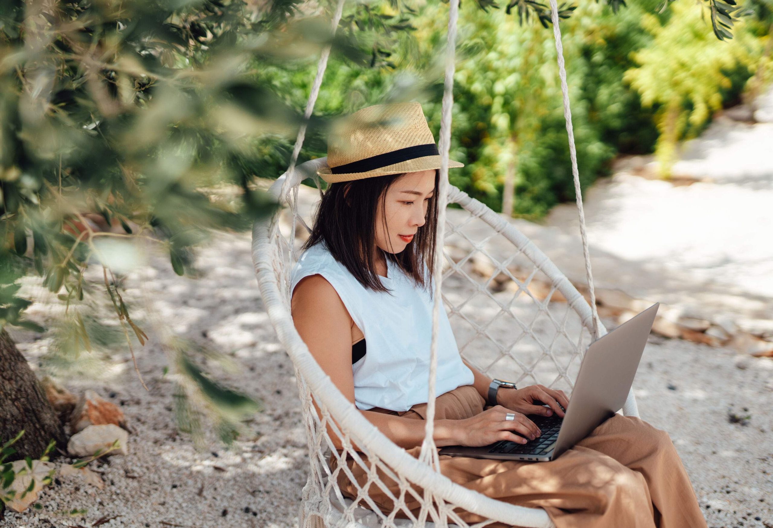 A woman working on her laptop while sitting on a swing chair hanging from a tree on the beach.