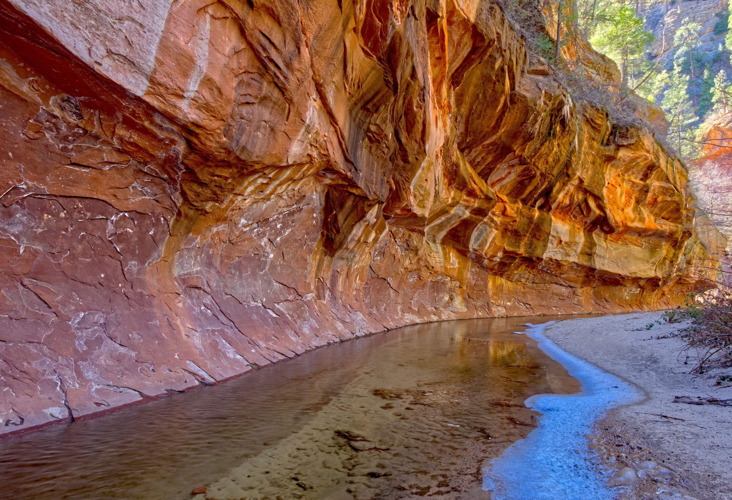 A rock wall with beautiful wavy carvings overhangs on a shallow stream.