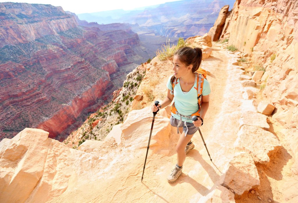A woman clutching trekking poles walking on a narrow path carved into the rocky mountains of the Grand Canyon, Arizona, USA.
