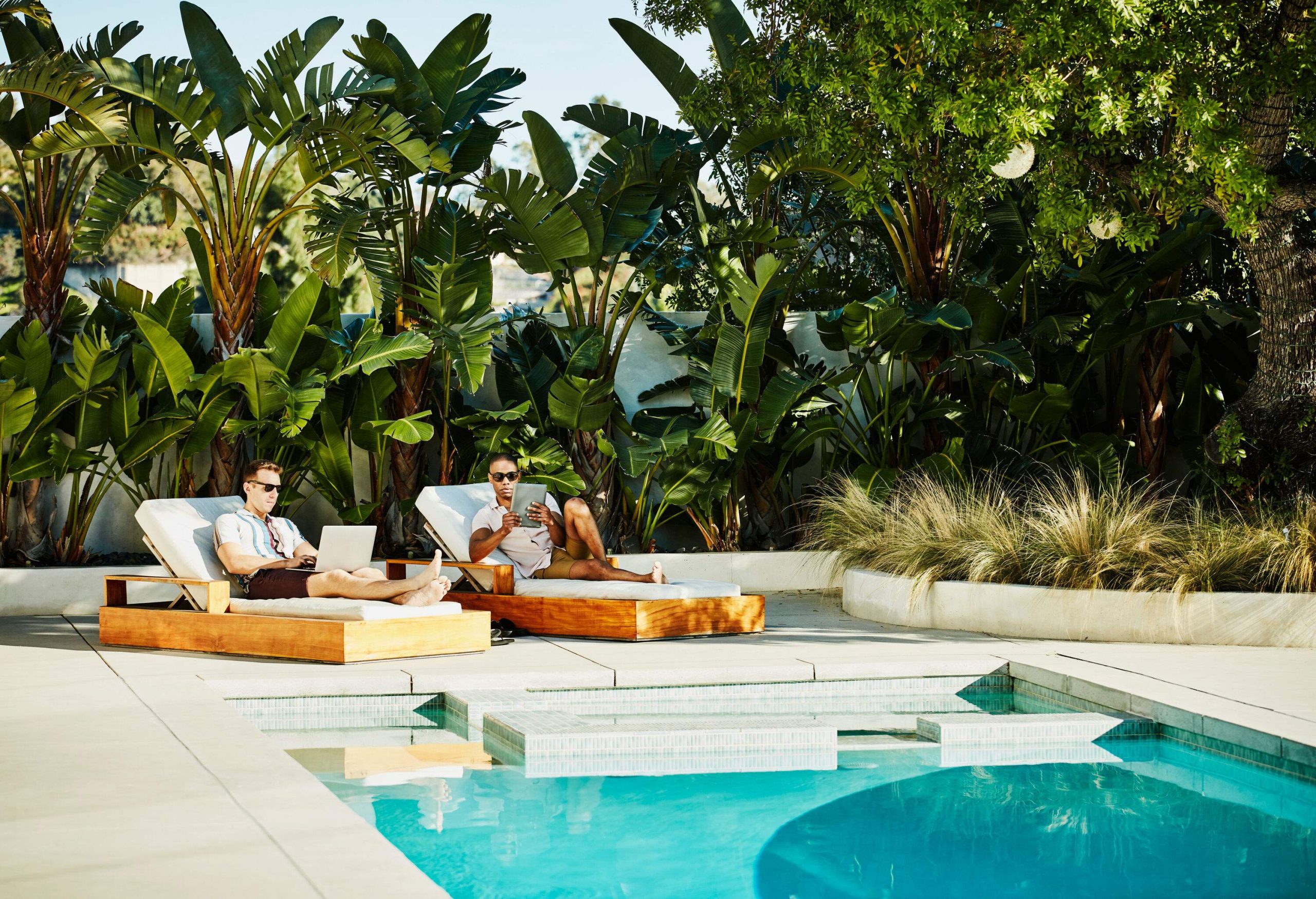 A gay couple on the sunloungers using their laptop and tablet on the poolside.