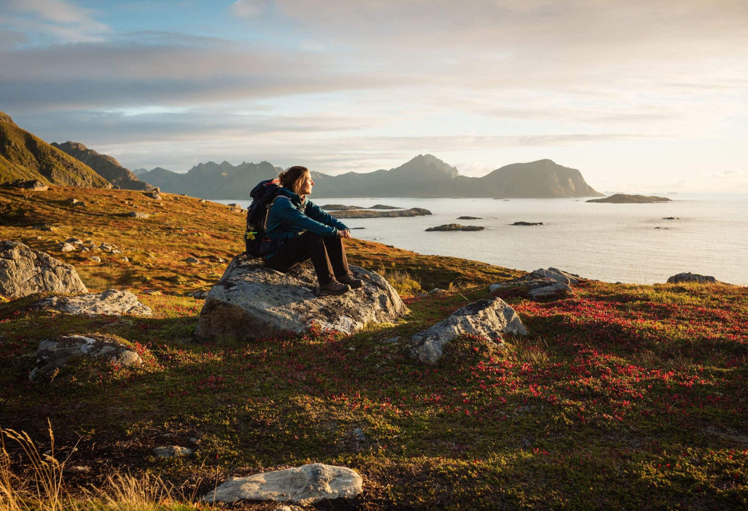 A woman in hiking gear sits on a boulder and gazes at a vast ocean.