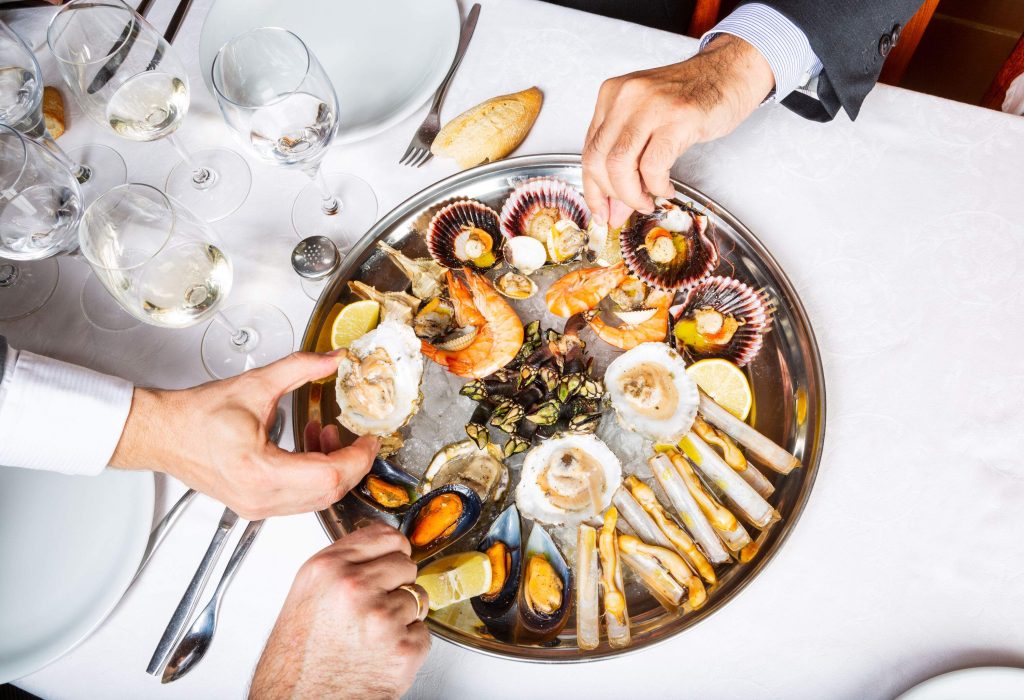 Several hands picking food from a stainless steel tray of finger foods that includes mussels, clams, and oysters.