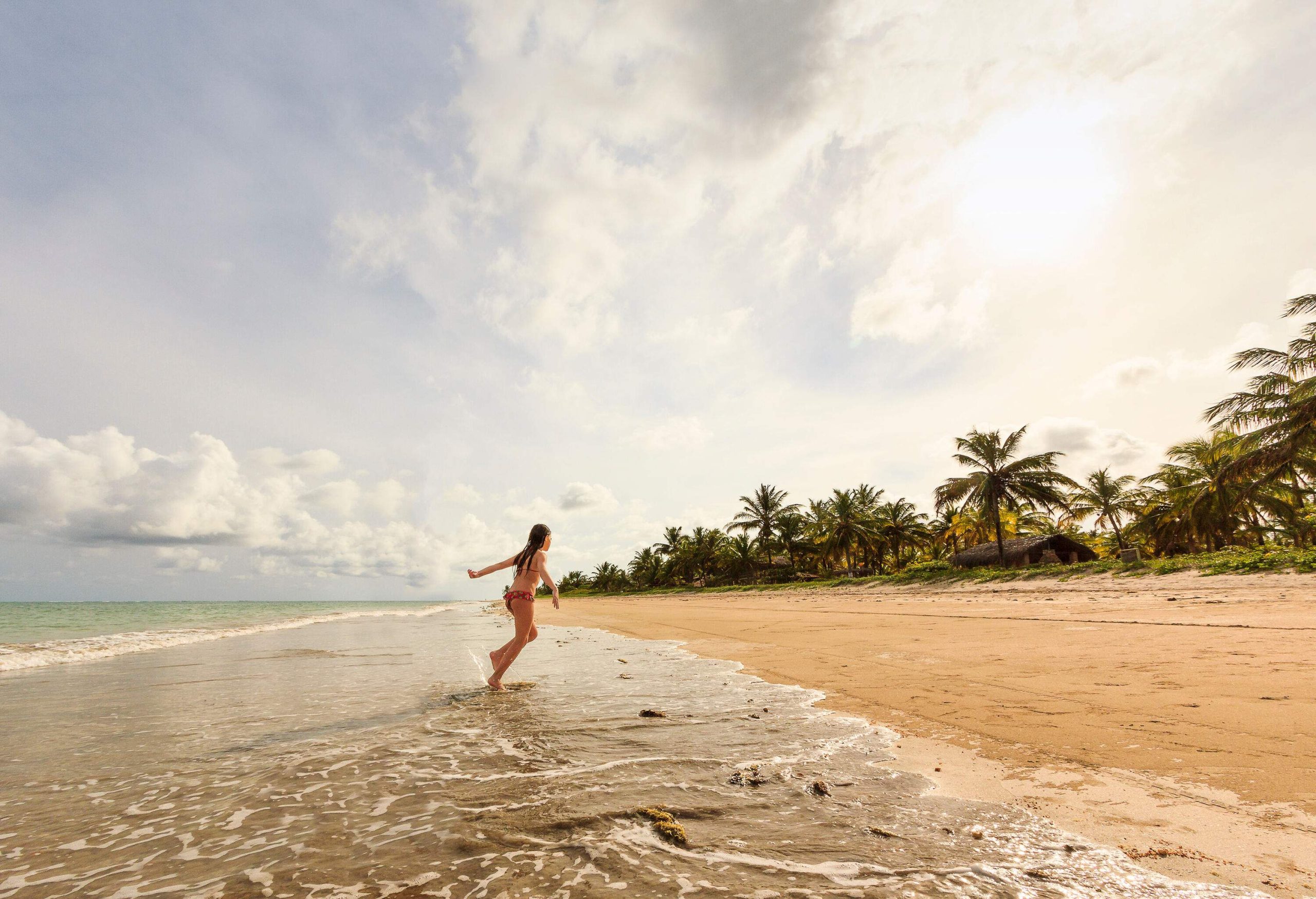 A person in a bikini joyfully runs towards the sandy beach from the water, with soft waves gently lapping the shore and tropical trees adorning the shoreline.
