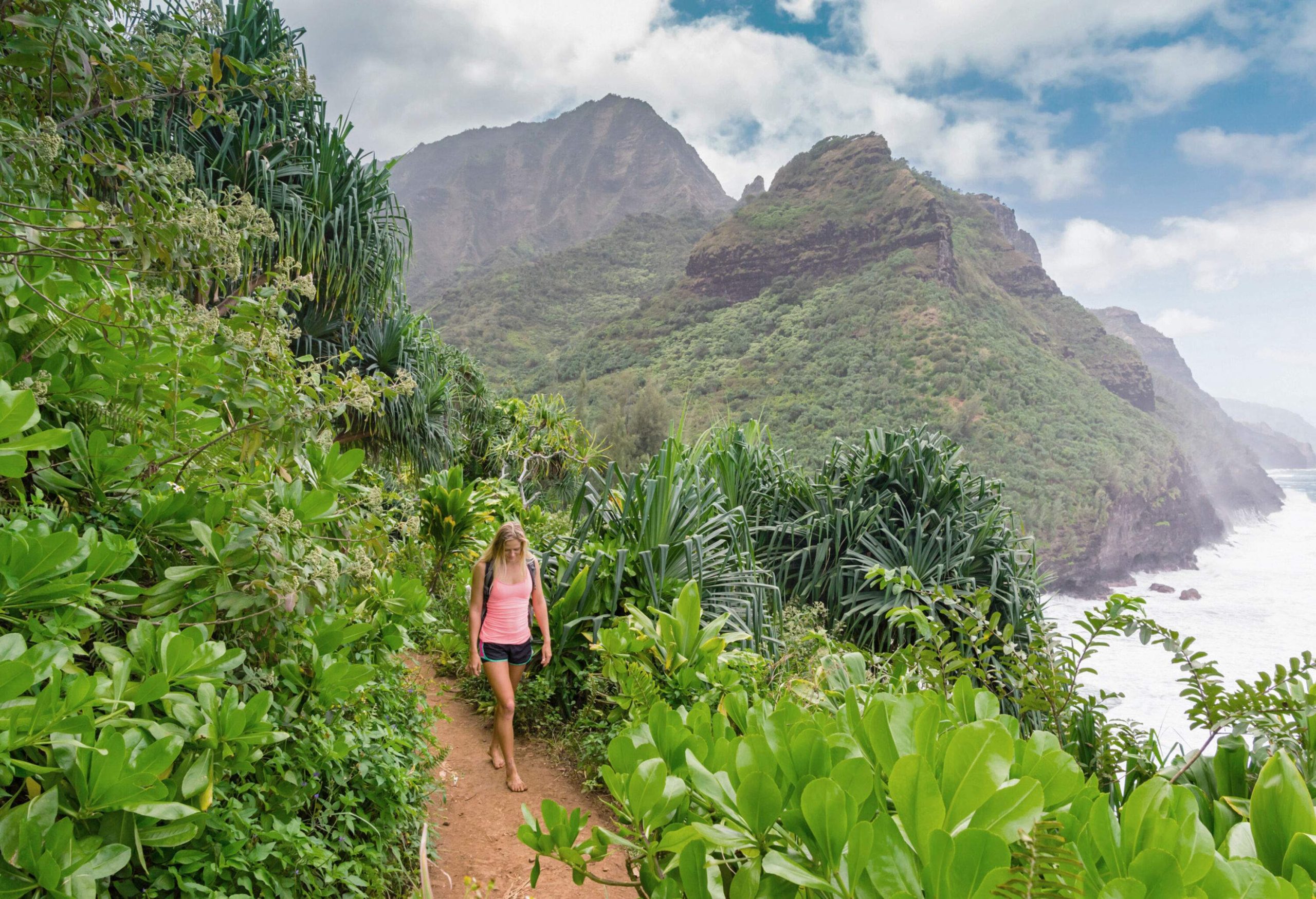 Woman hiking barefoot in a forest on a hillside trail with views of the ocean crashing to the distant mountain coast.