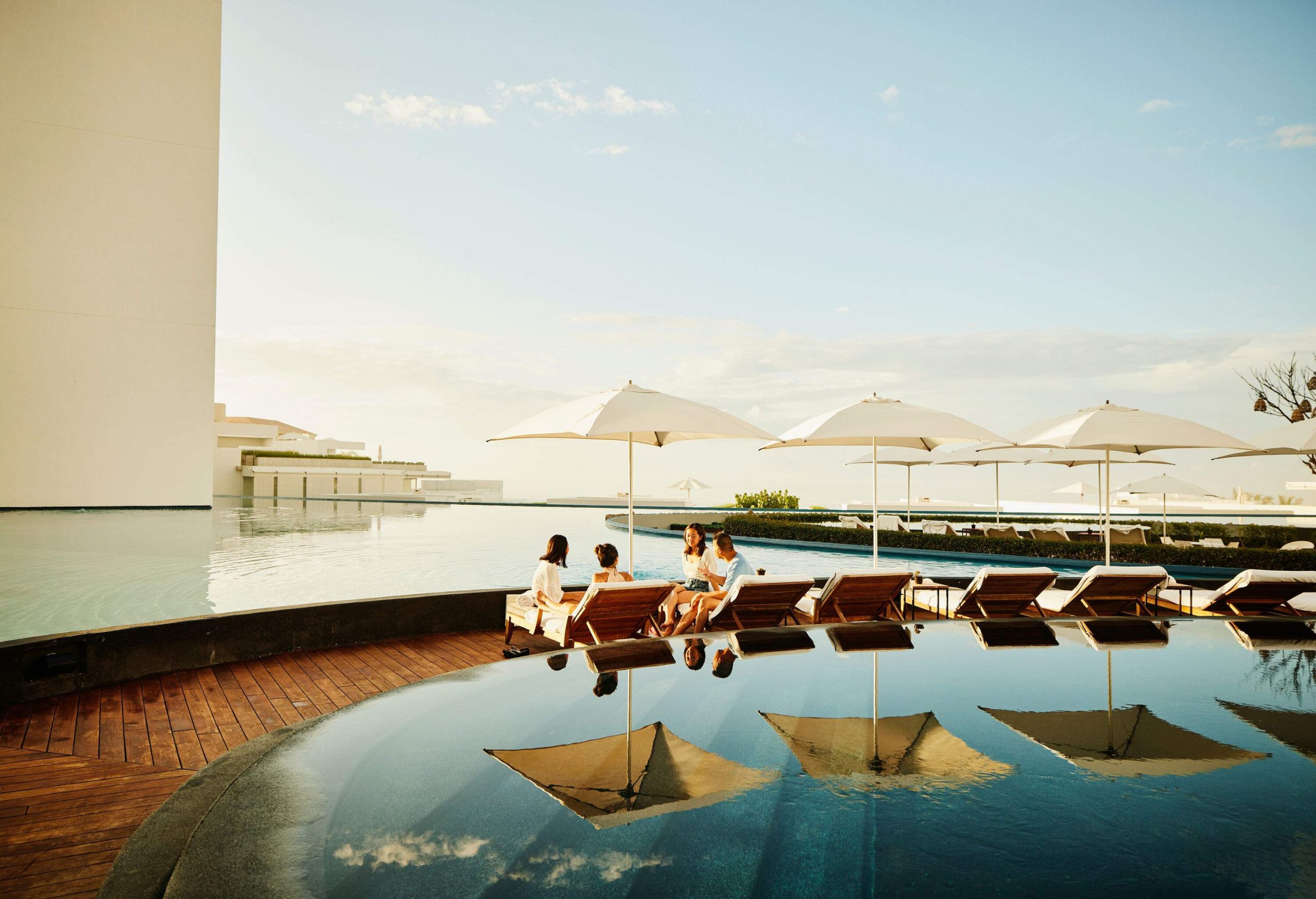 A circle of friends having a good conversation while sitting on the wooden sun loungers at the poolside.