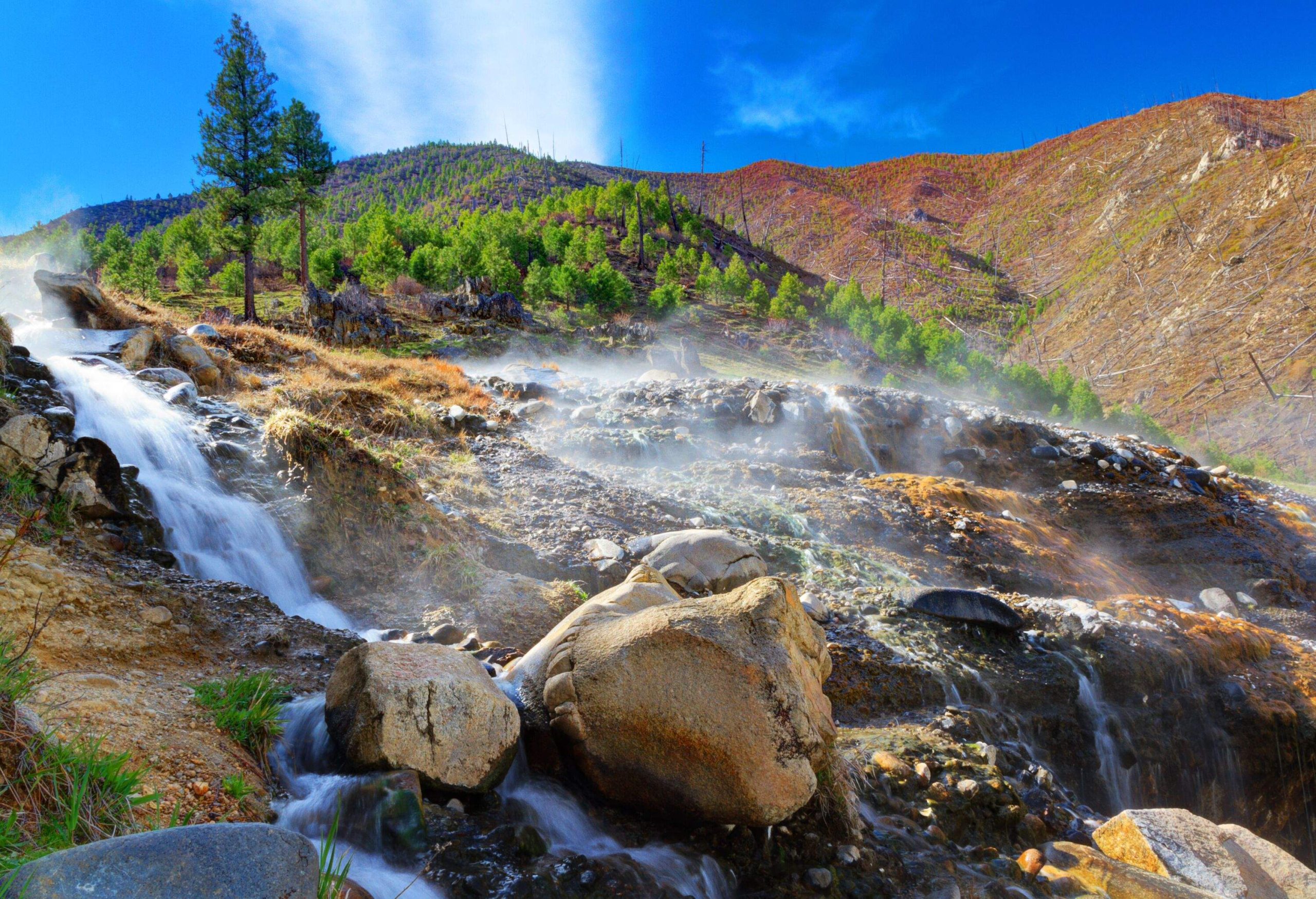 Small steaming waterfalls pour down hillside to feed Payette River on partly cloudy spring morning at Kirkham Hot Springs outside Lowman, Idaho