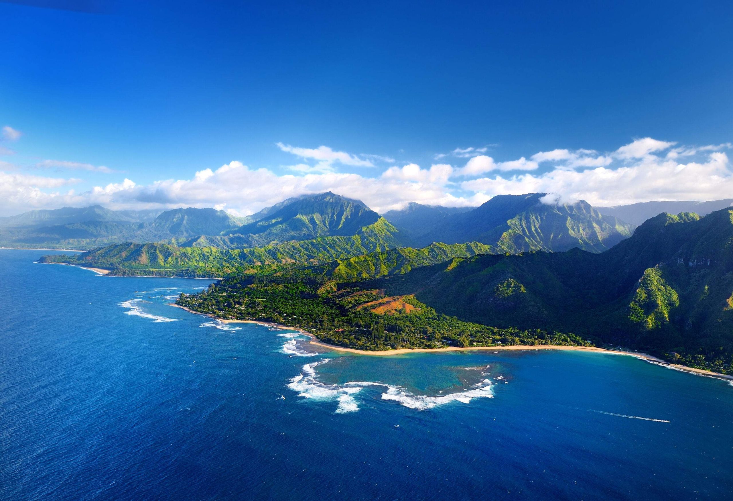 Calm blue sea with small waves and the mountain range covered in lush vegetation against the cloudy blue sky.