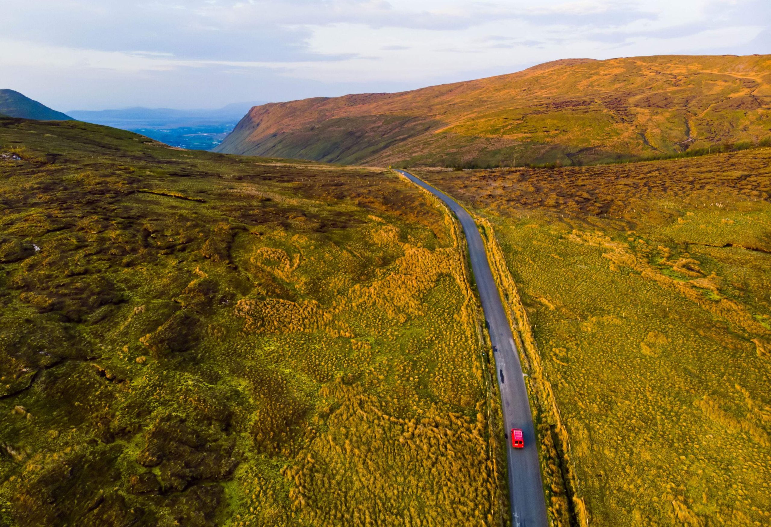 A red car driving down a road through a vast countryside with mountains in the distance.