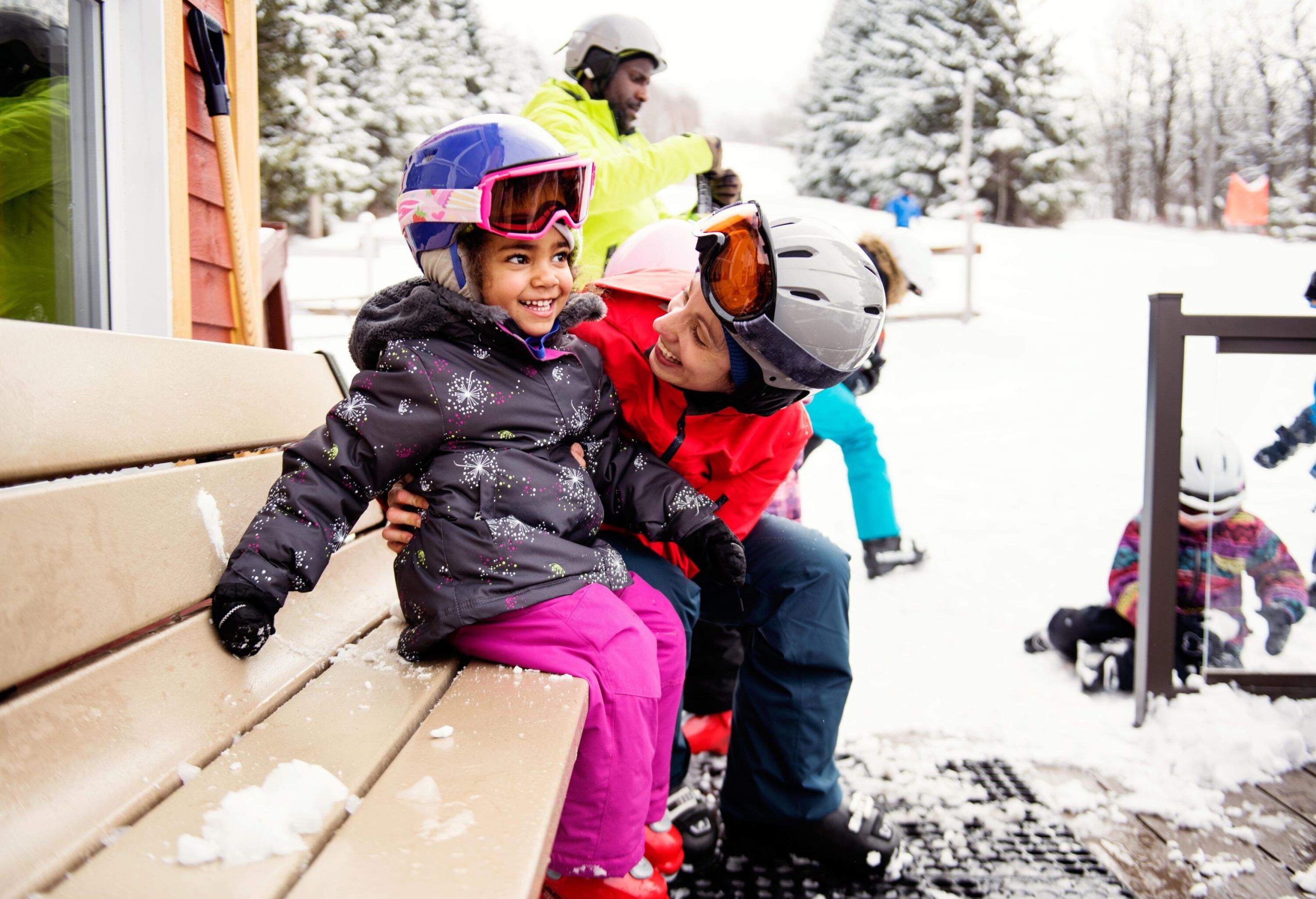 Joyful mom and child in colourful winter attire sitting next to skiers in the background.