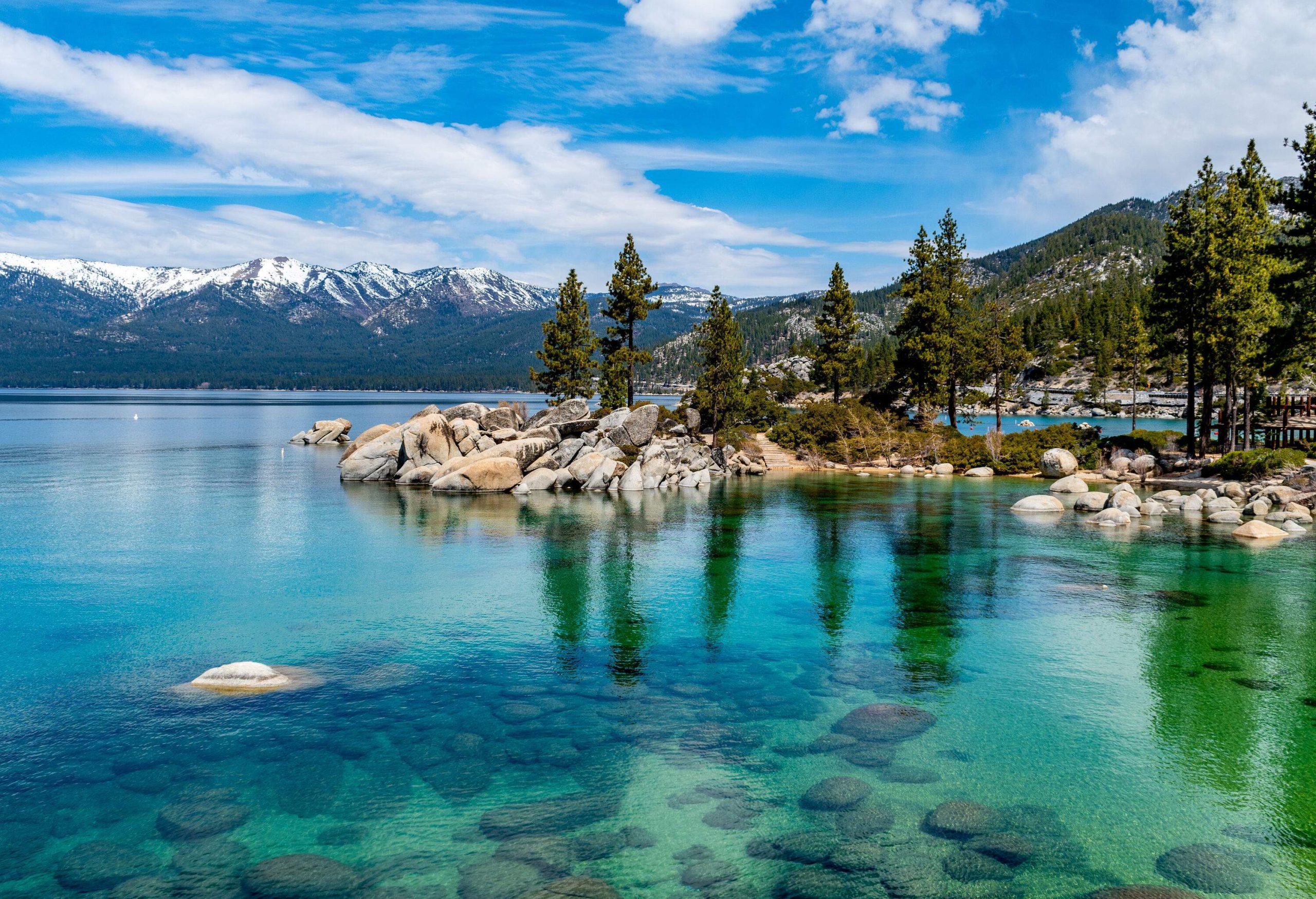 A tranquil clear water lake reflects the lined tall green trees among the boulders and the mountain range against the blue sky.