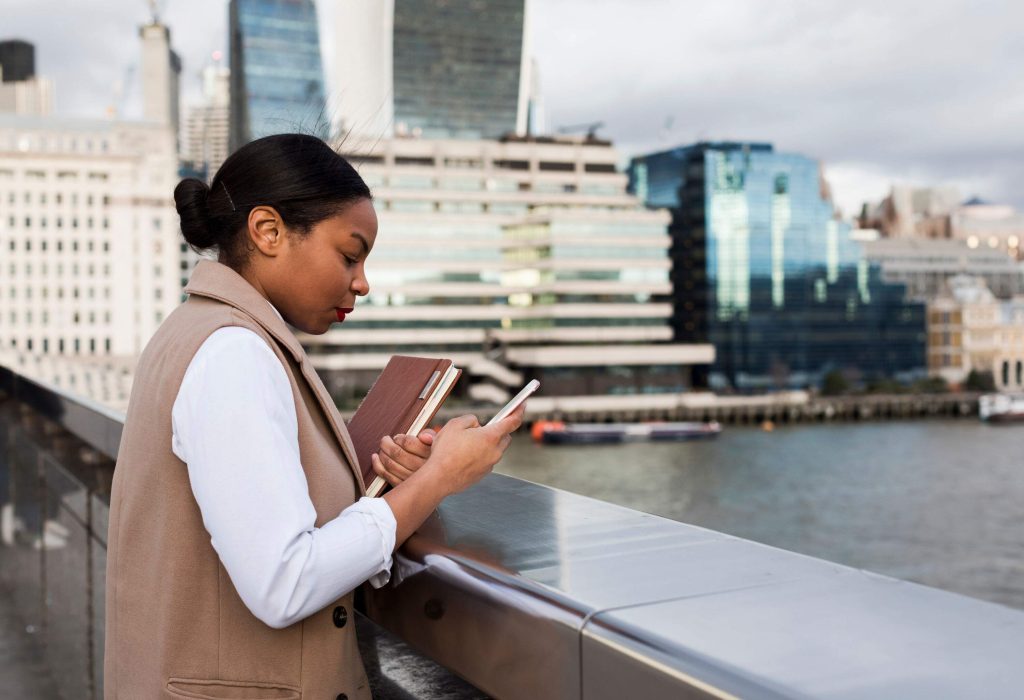 A woman standing on a bridge holding a book and using her smartphone.