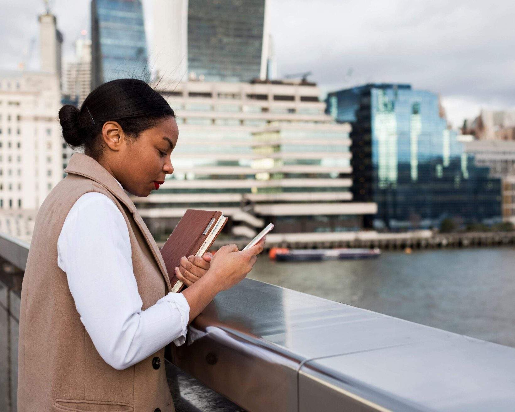 A woman standing on a bridge holding a book and using her smartphone.