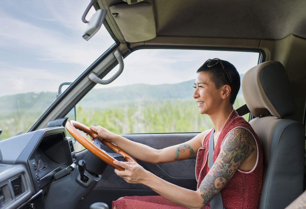 A smiling lady with tattoos and shaved hairstyle in a red dress drives the car.