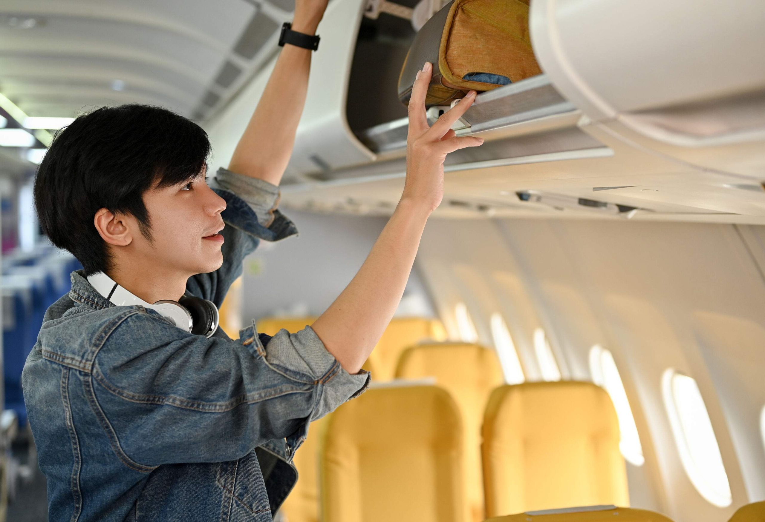 A young man stows his hand-carry luggage in the overhead compartment of an aircraft.