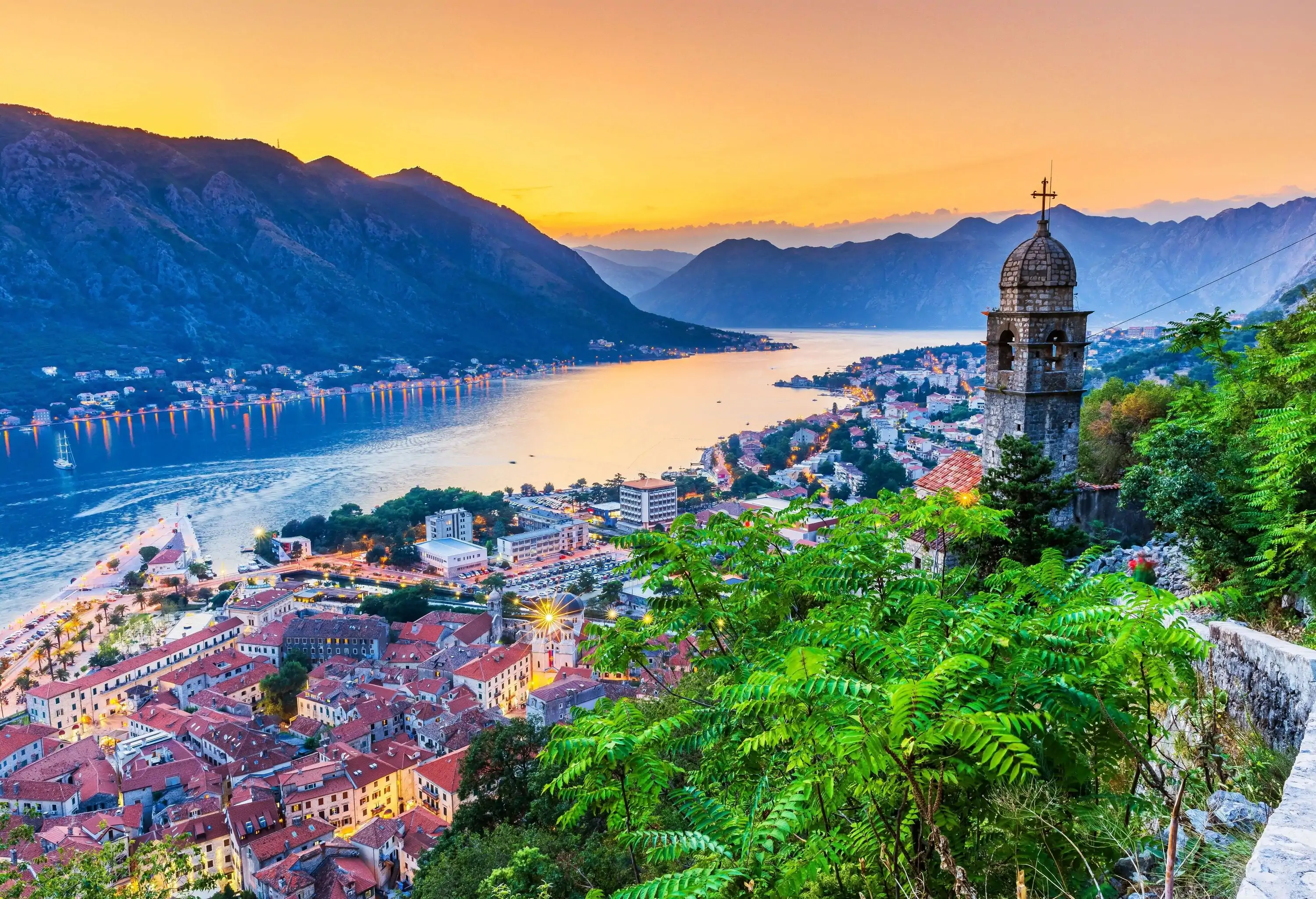 Scenic aerial view of an old seaside town with a Romanesque church bell tower overlooking the mountains across the bay against a dramatic twilight sky.