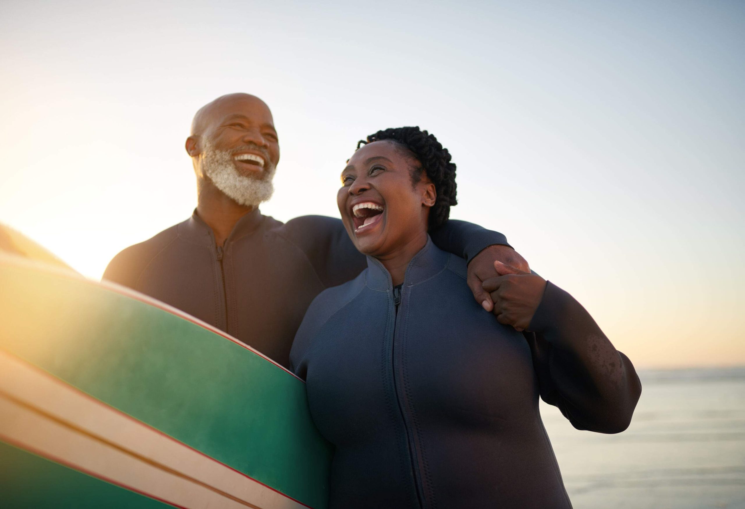 A couple laughing together while carrying surfboards on the beach.