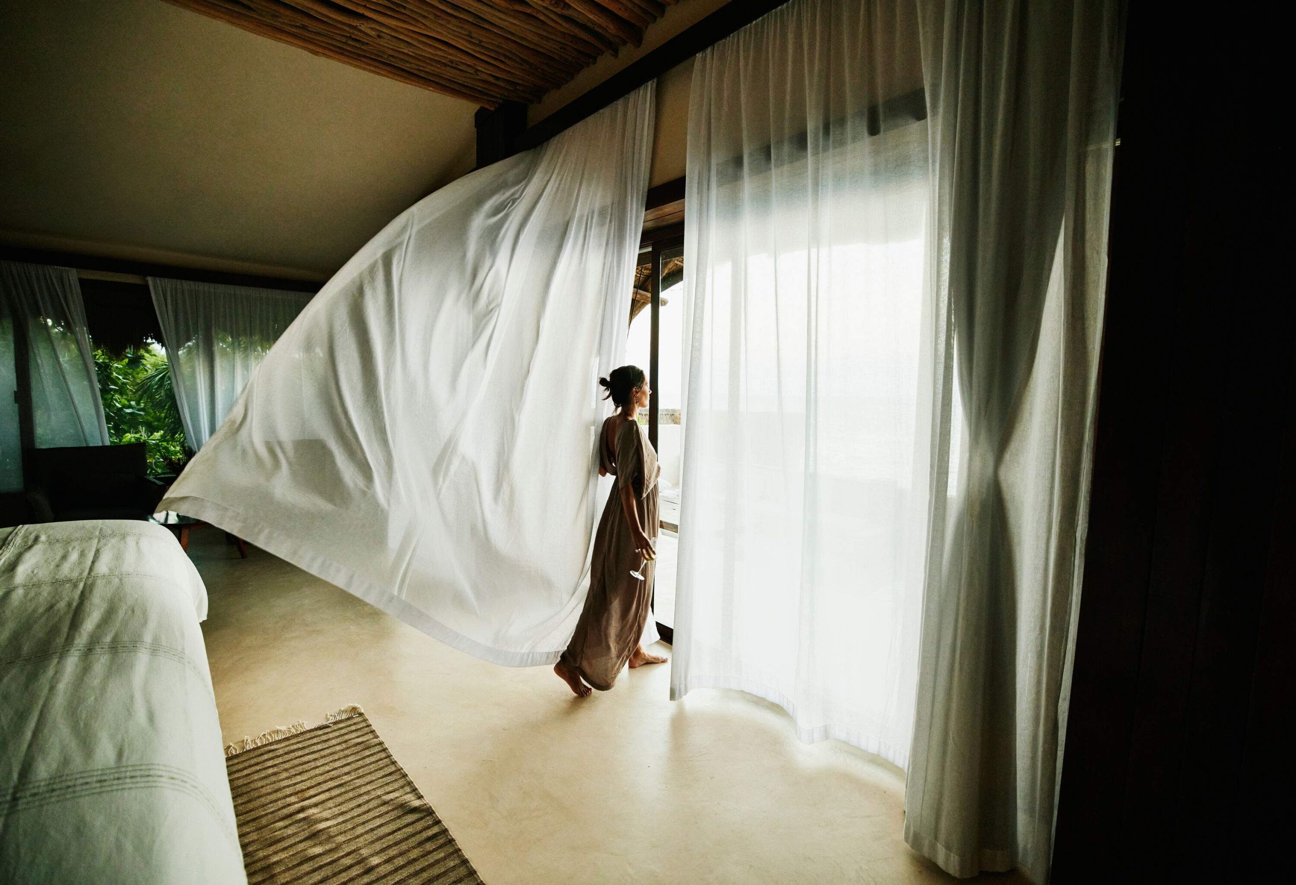 A woman stands beside a big window as the breeze flows through the curtains.