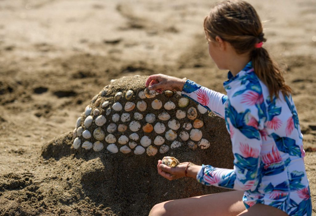 A girl covers the sandhill with seashells on the beach.