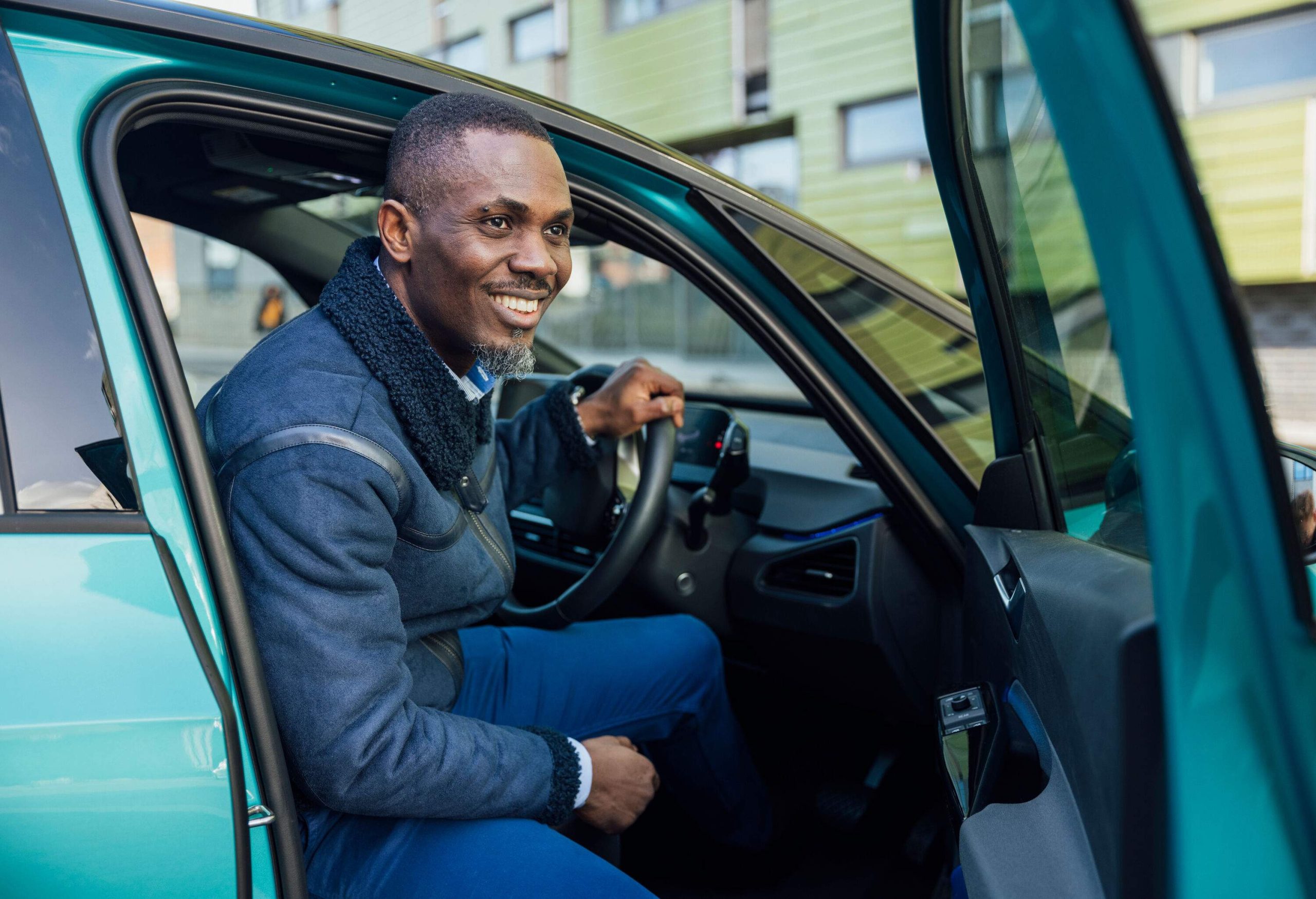 A smiling, dark-skinned man coming out of his blue four-wheel vehicle.
